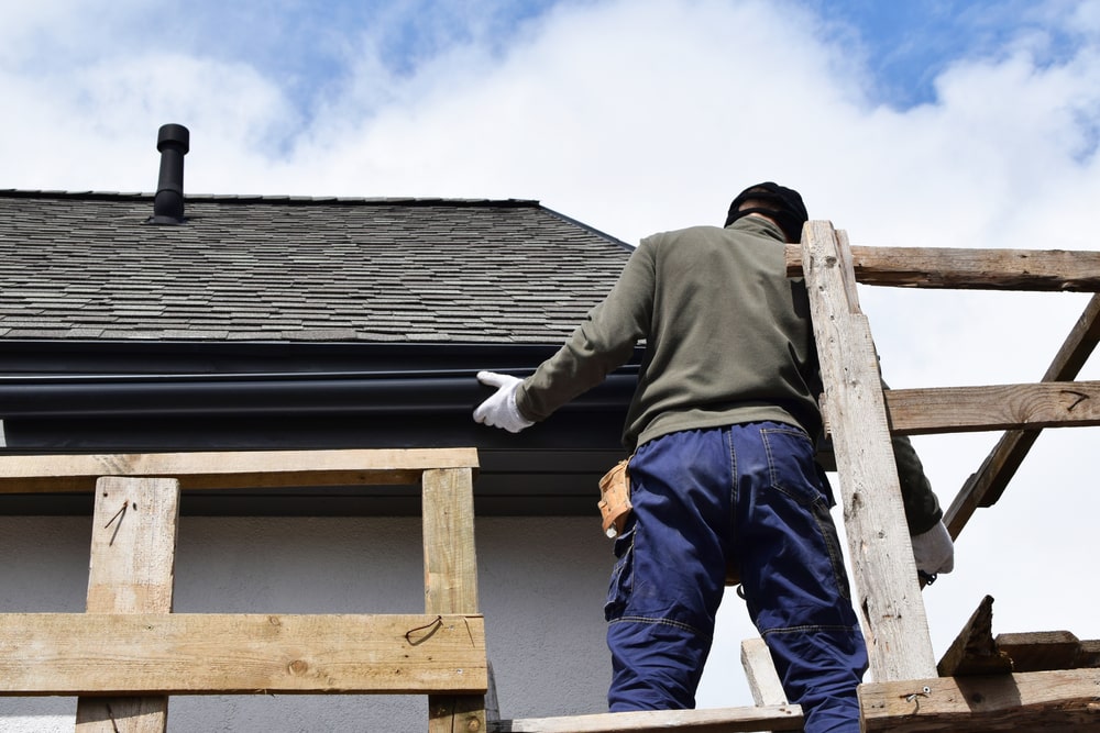 Construction Worker Installing Plastic Roof Rain Gutter Gray Shingles Roof.