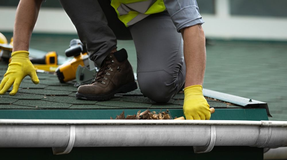 A Man Worker Is Cleaning Leaves From A Clogged Roof Gutter