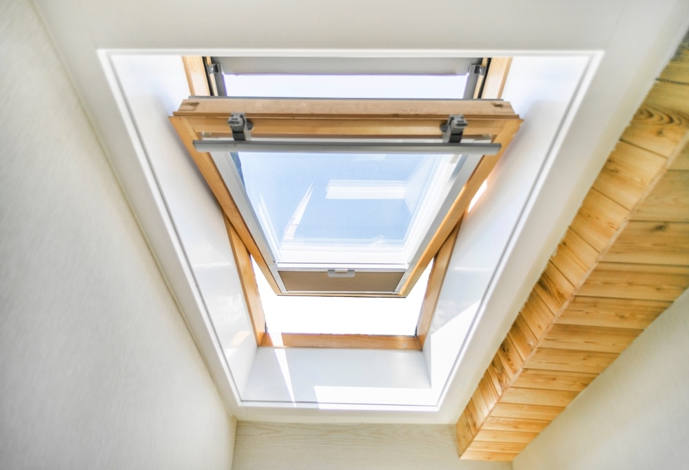 A wooden-framed skylight window is open, allowing sunlight to enter. Wooden steps lead up to the window, and the sky is visible through the glass. Perfect inspiration for any home improvement Suffolk County, NY project.