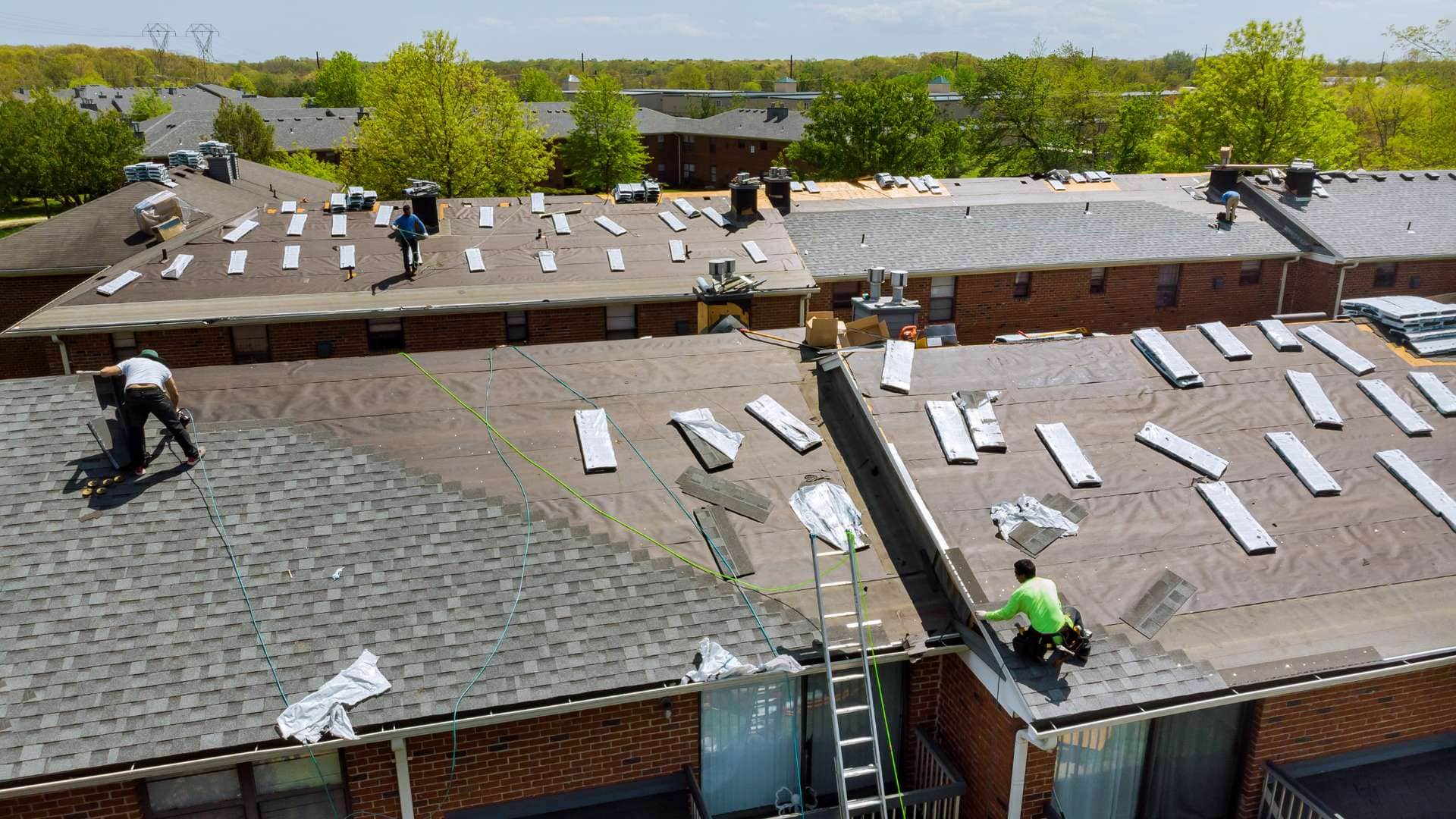 Aerial view of a roofing contractor in Suffolk County as workers install new shingles on apartment buildings; bundles of materials are spaced across the rooftops, and safety ropes secure the crew during this home improvement in NY.