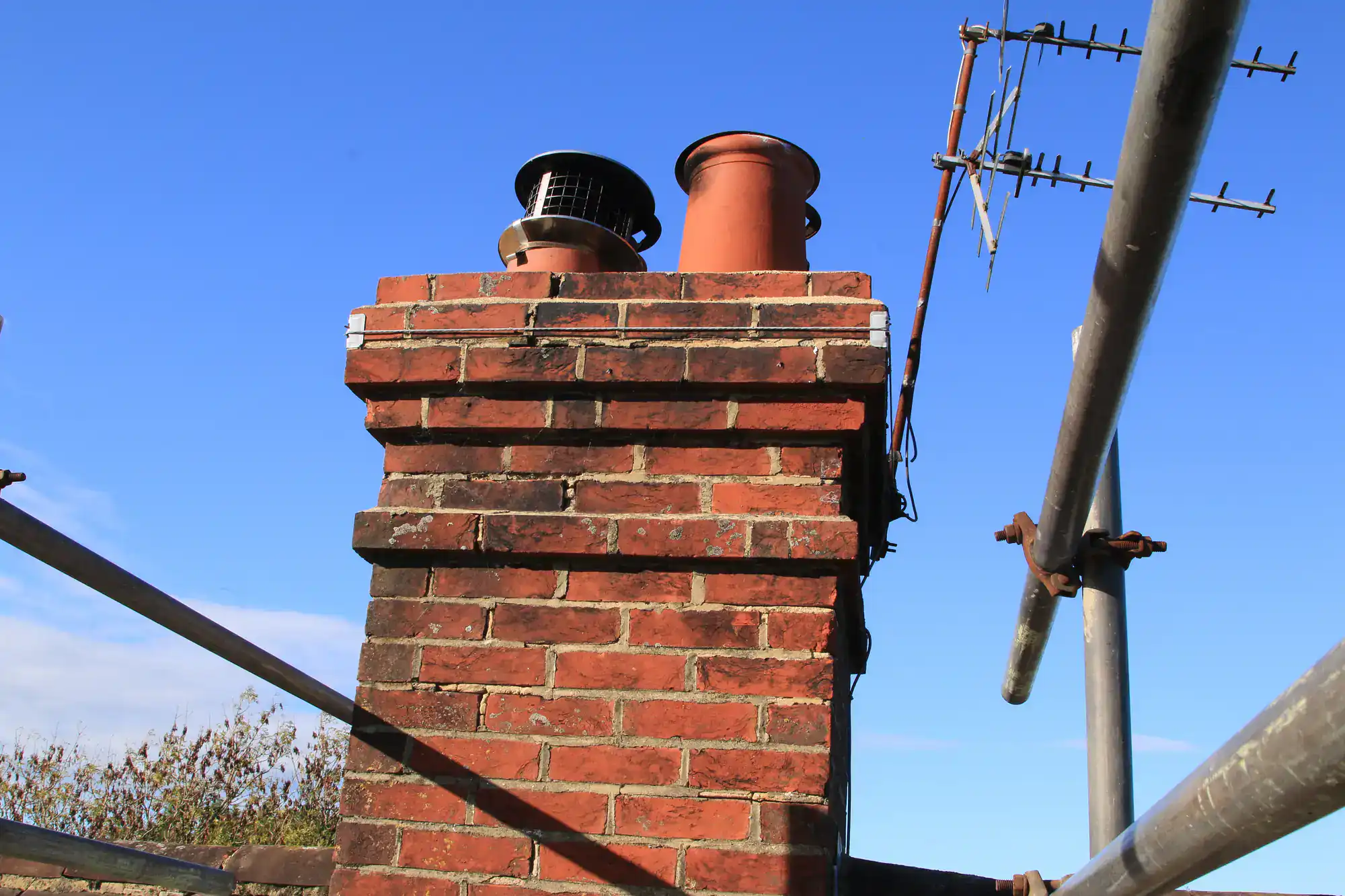 A red brick chimney with two pots—one metal and one terracotta—viewed from below, surrounded by scaffolding poles, with a TV aerial attached and a clear blue sky in the background.
