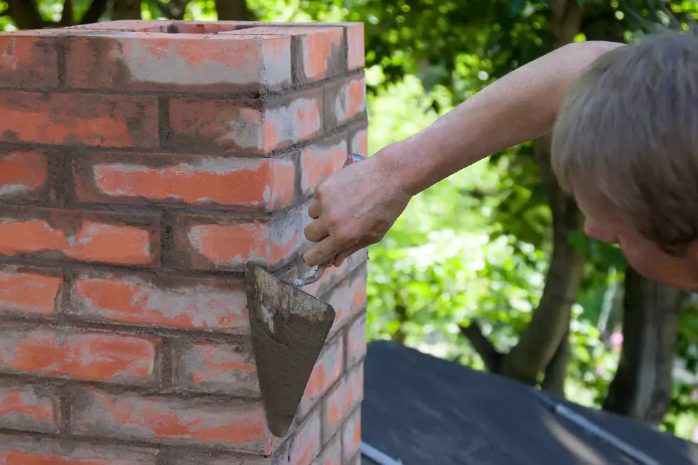 A person uses a trowel to apply mortar to bricks while constructing or repairing a brick chimney outdoors, surrounded by green trees.