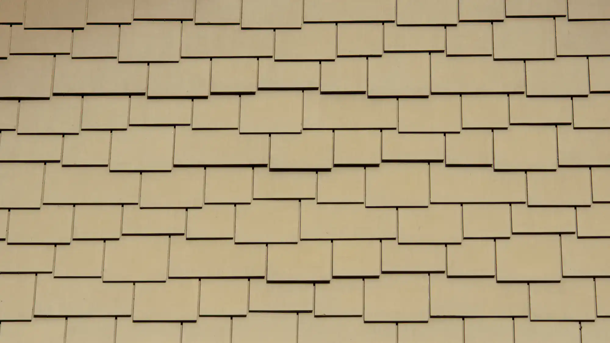 A close-up view of a beige shingle wall with overlapping rectangular tiles arranged in a staggered, uniform pattern.