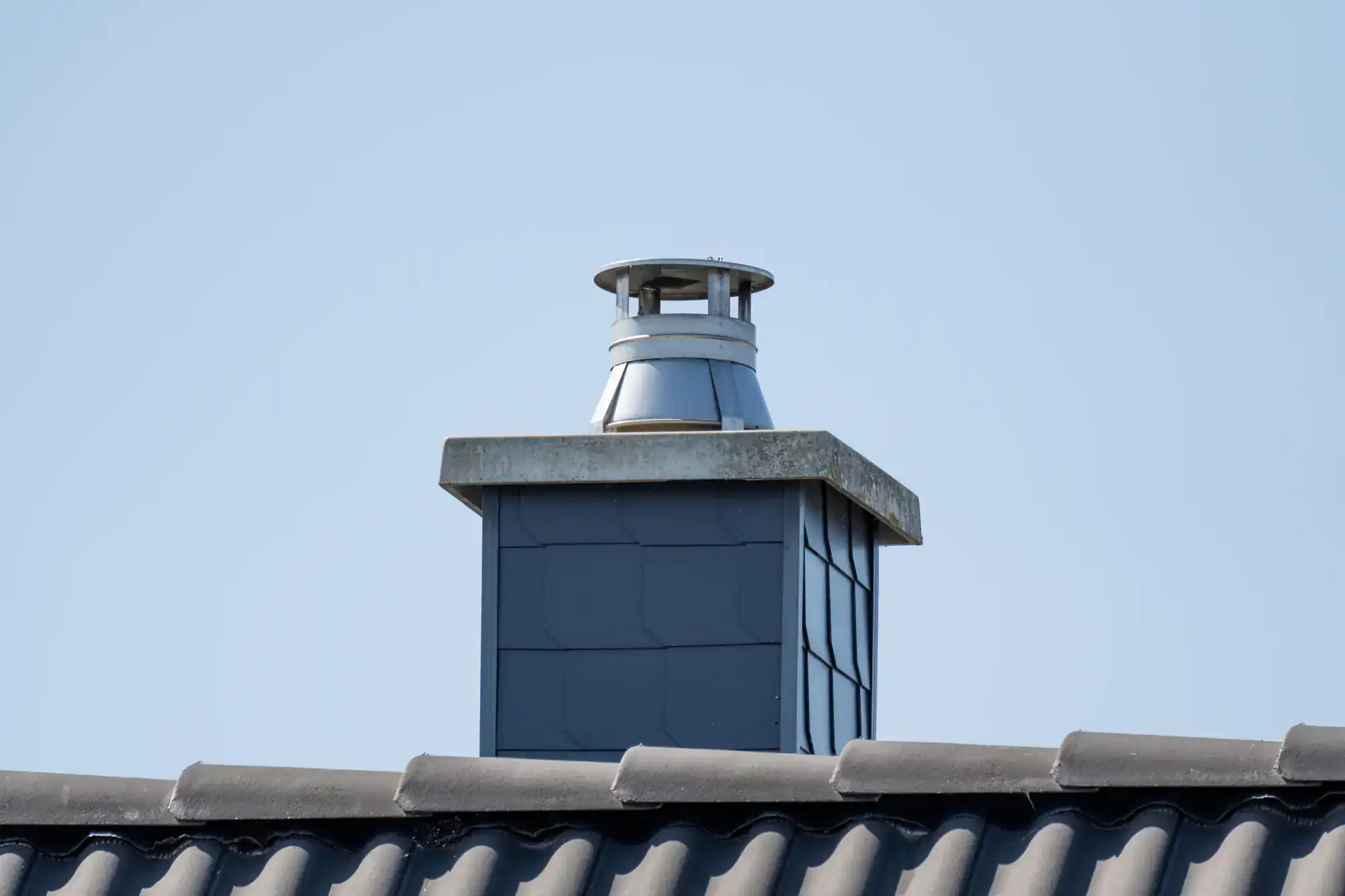 A metal chimney cap sits on top of a square chimney with dark tiles, extending above a dark, wavy tiled roof against a clear blue sky.