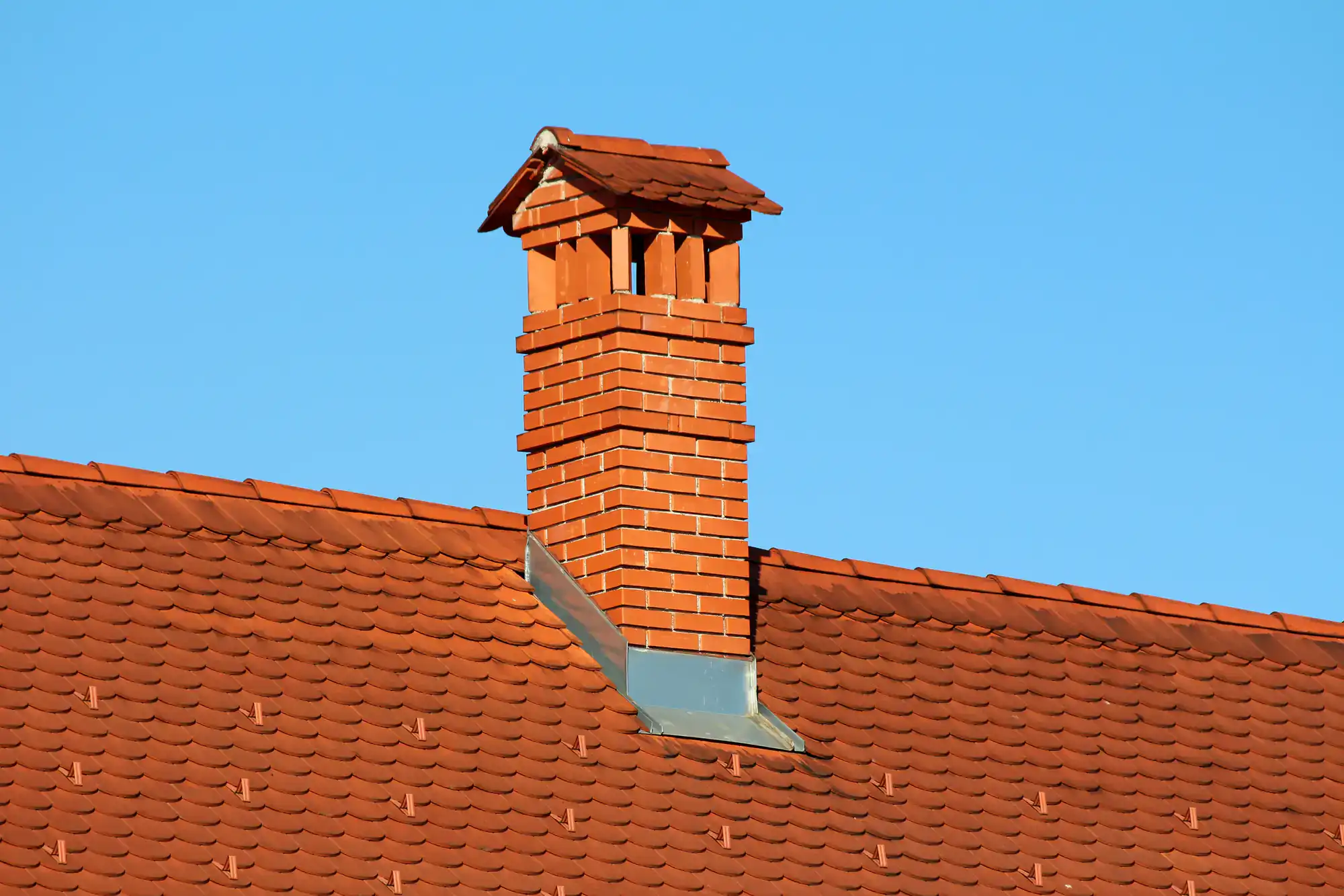 A red brick chimney with a small tiled roof sits atop a sloped roof covered in red tiles, set against a clear blue sky.