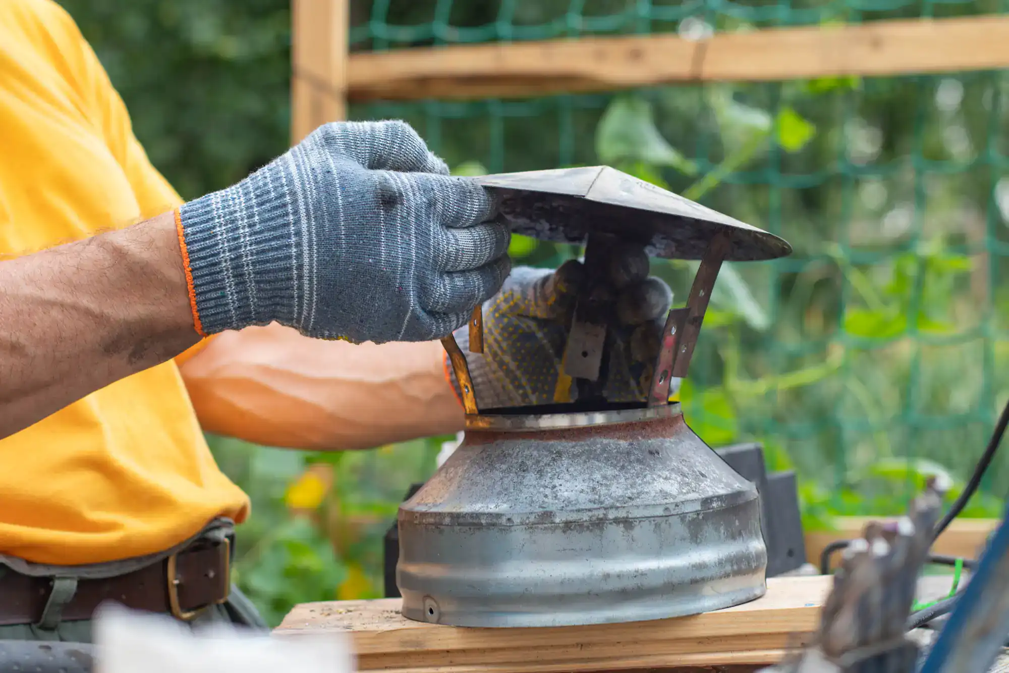 A person wearing work gloves and a yellow shirt repairs or assembles a metal outdoor lantern on a wooden workbench, with greenery and a wooden structure in the background.