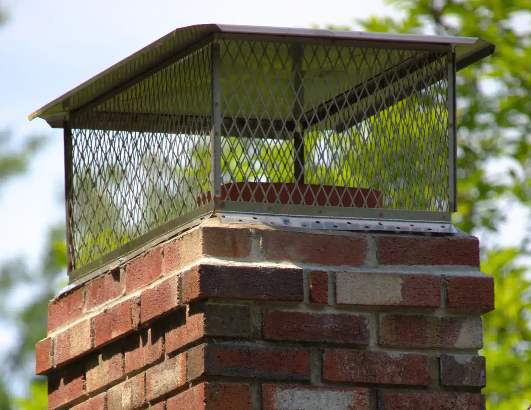 A close-up of a brick chimney with a metal wire mesh cap on top, set against a background of green trees and blue sky. The metal cap is designed to prevent debris or animals from entering the chimney.
