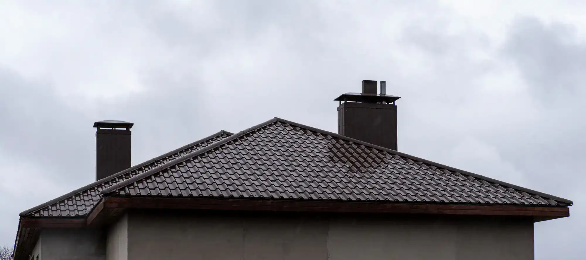 A house with a dark, tiled, sloped roof and two chimneys, set against a cloudy, overcast sky.