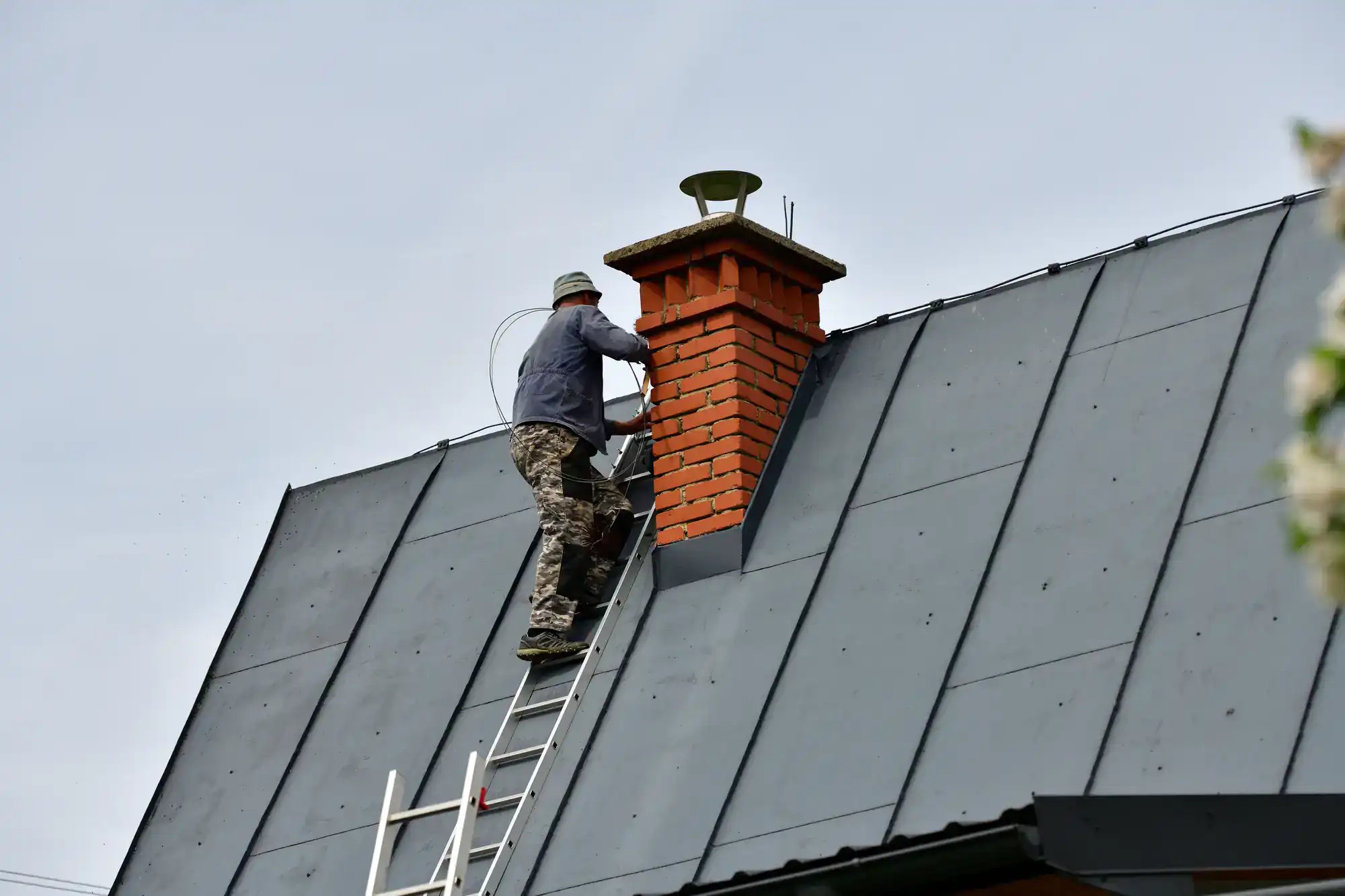 A person in camouflage pants and a hat stands on a ladder leaning against a steep metal roof, working on a brick chimney under a cloudy sky.