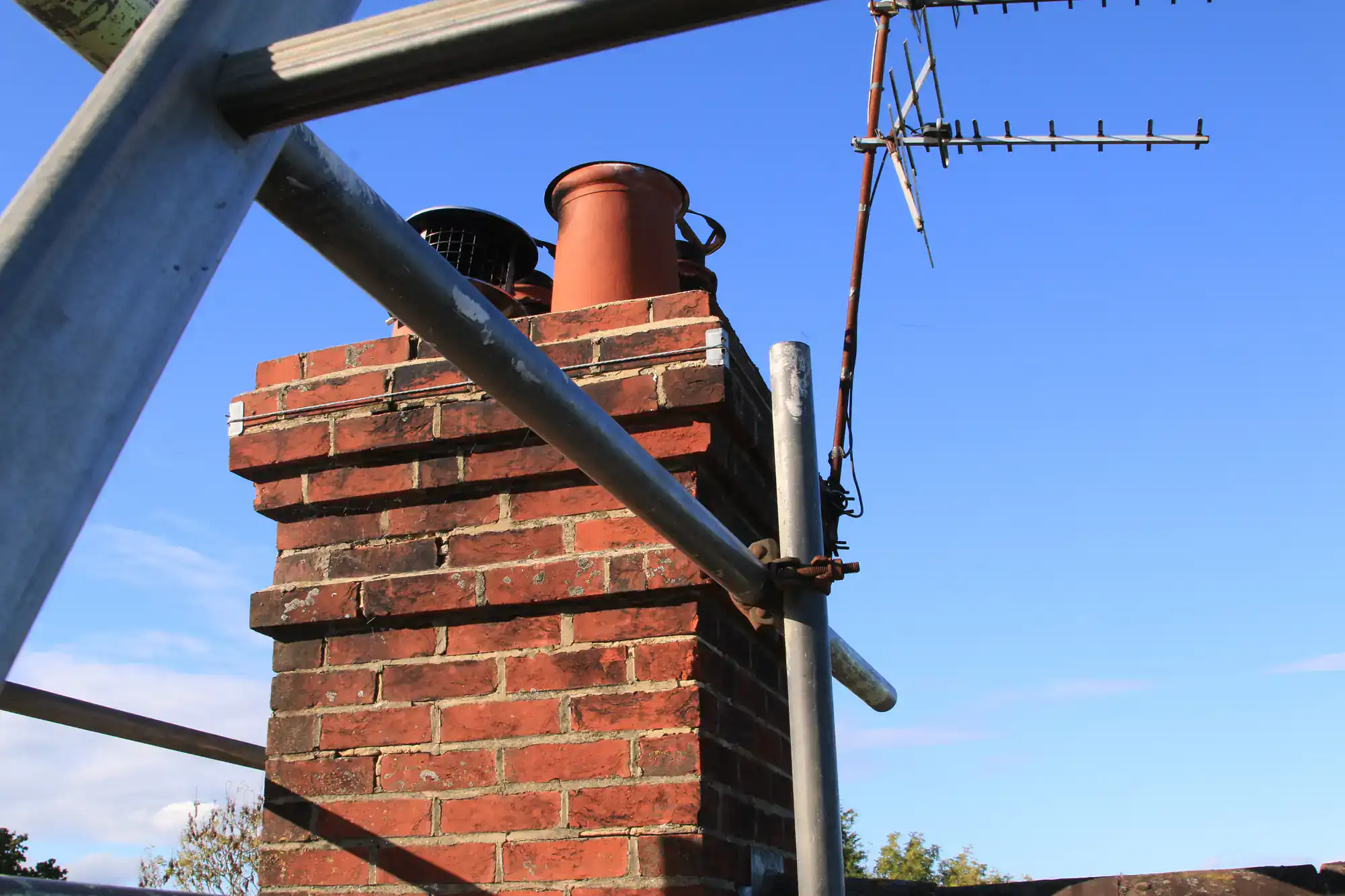 A close-up of a red brick chimney with two chimney pots and a TV antenna, surrounded by metal scaffolding under a clear blue sky.