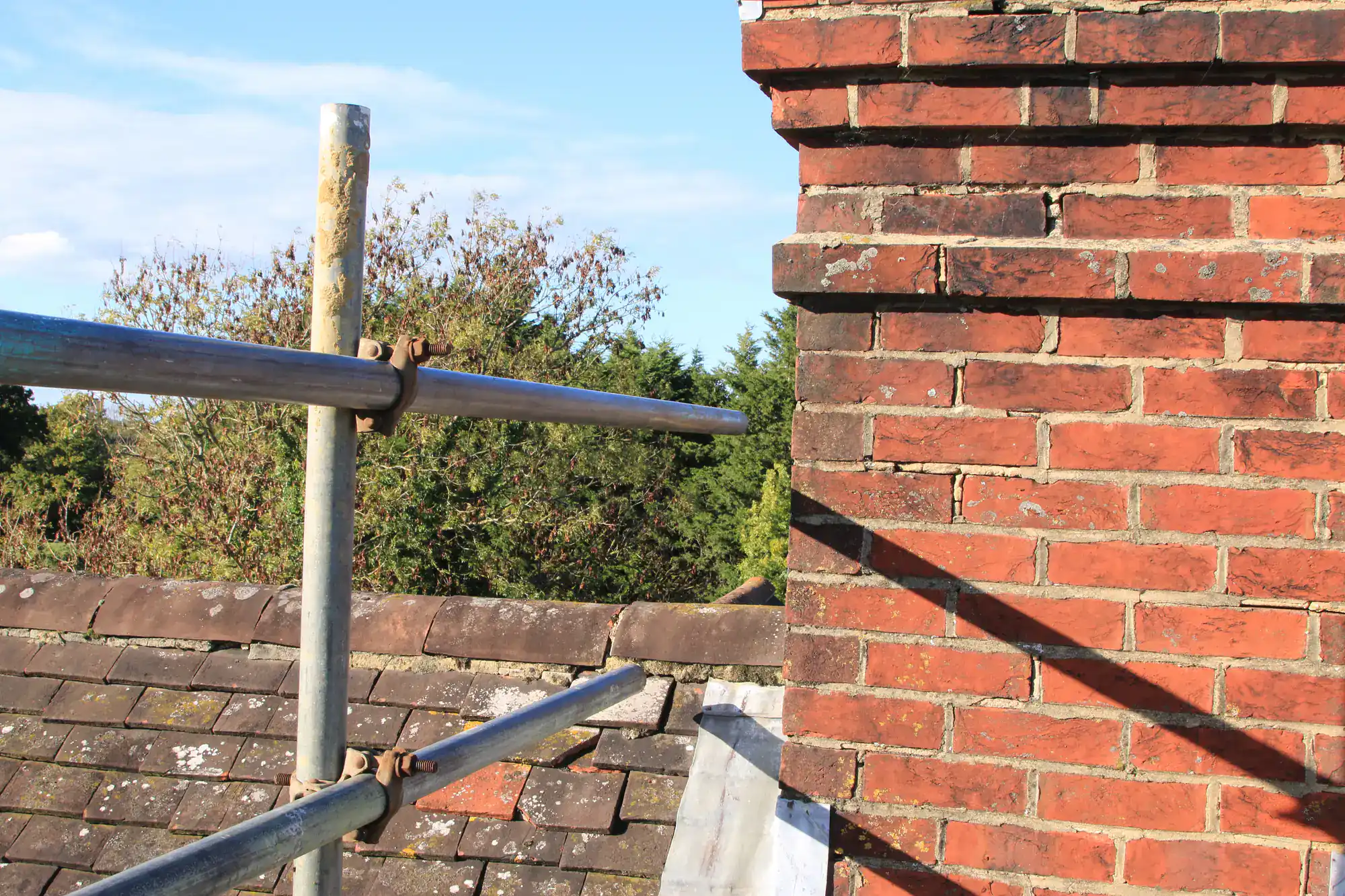 A close-up of a red brick chimney on a tiled roof, with metal scaffolding poles in front and trees in the background under a blue sky.