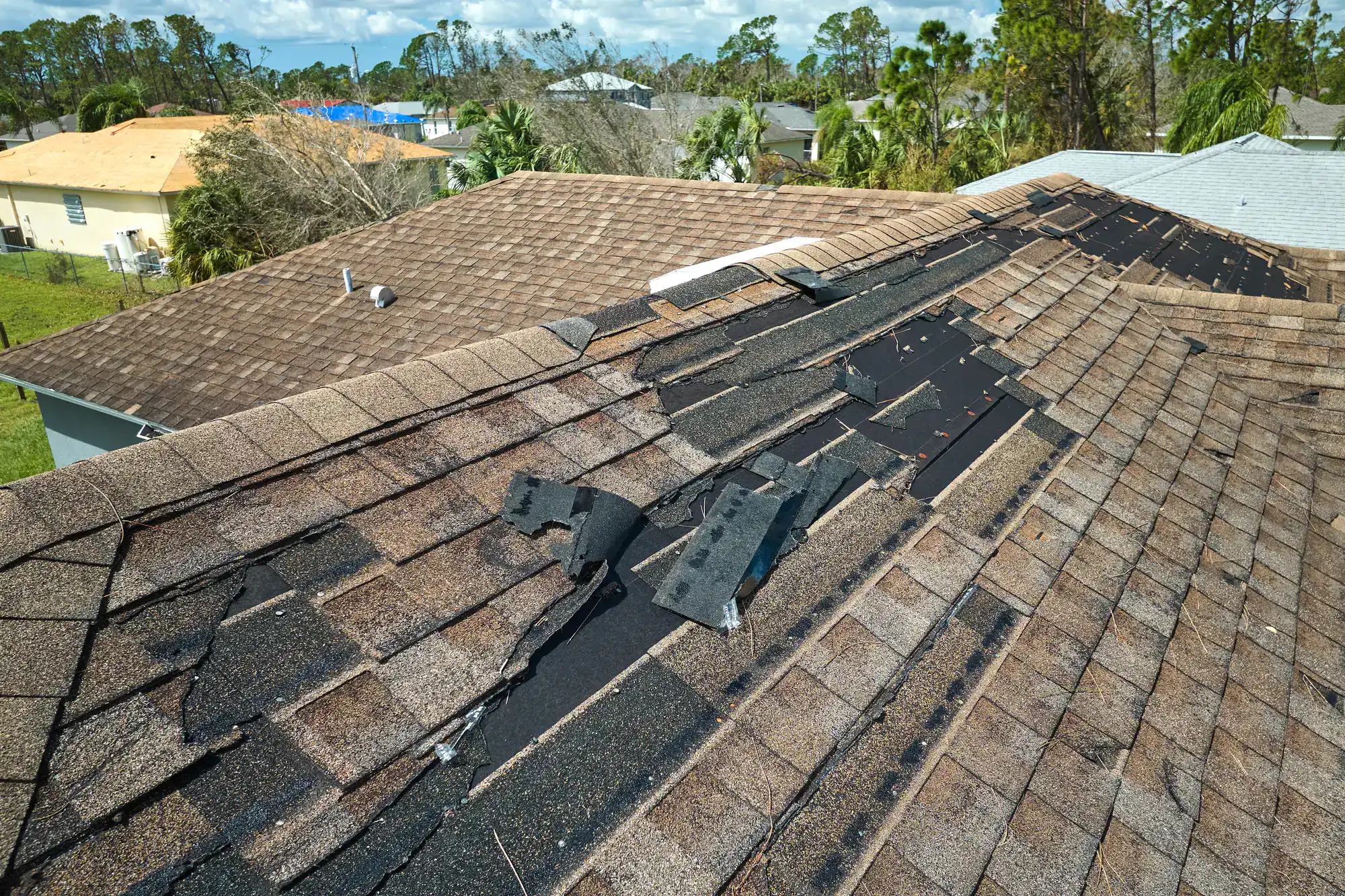 A house roof with missing and damaged shingles, exposing black underlayment beneath. Nearby roofs appear intact. Surrounding area has trees and other houses, with partly cloudy skies above.