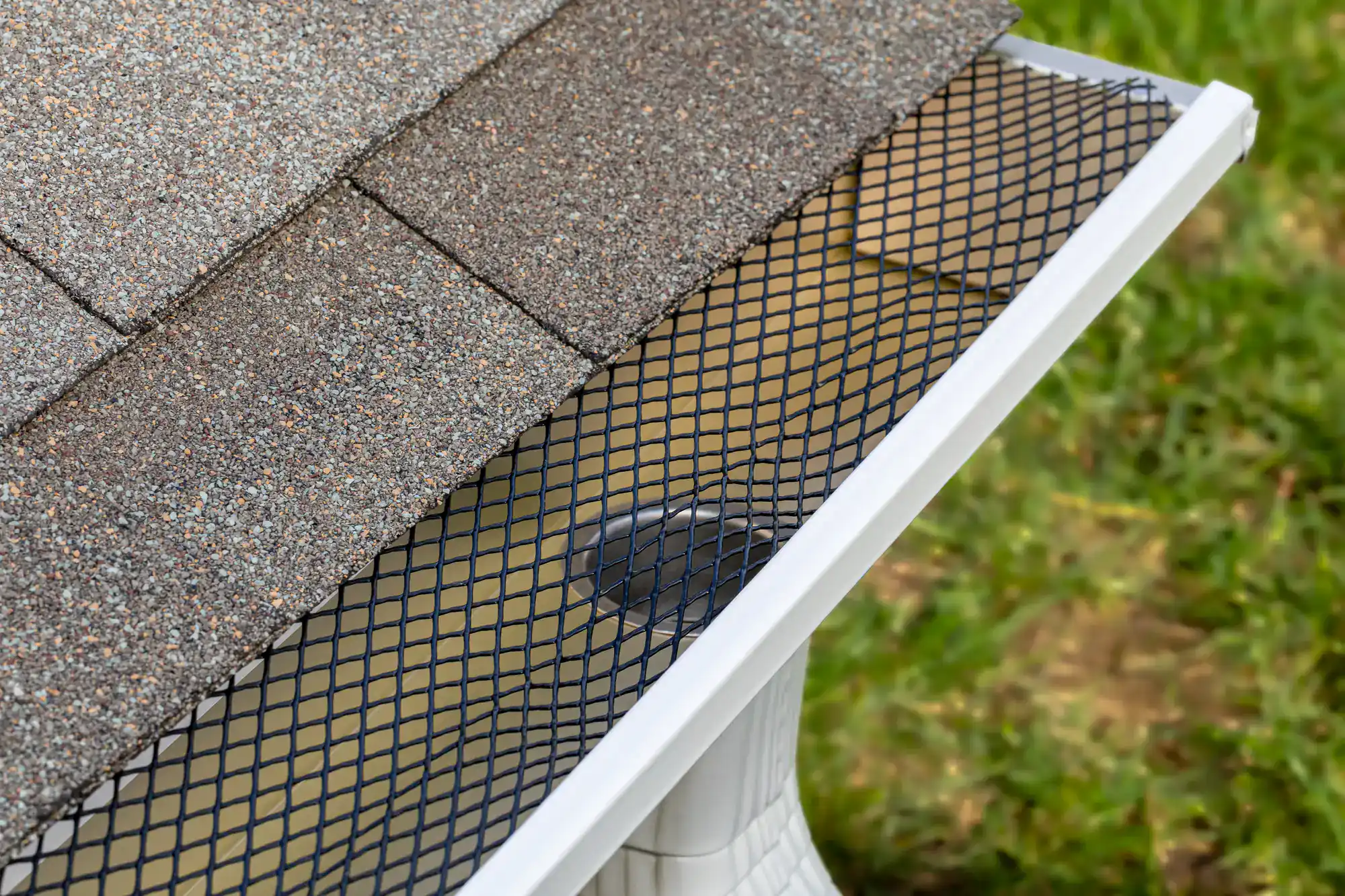 Close-up of a house roof’s edge showing a black mesh gutter guard installed over a white rain gutter, with brown shingles above and green grass visible in the background.