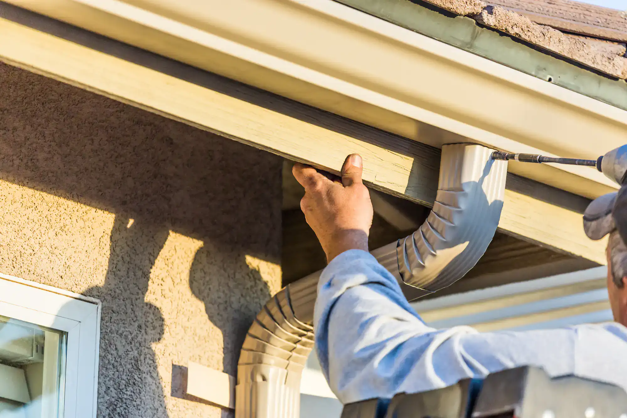 A person on a ladder installs or repairs a residential rain gutter system, securing the metal gutter to the roof edge of a house in sunlight.