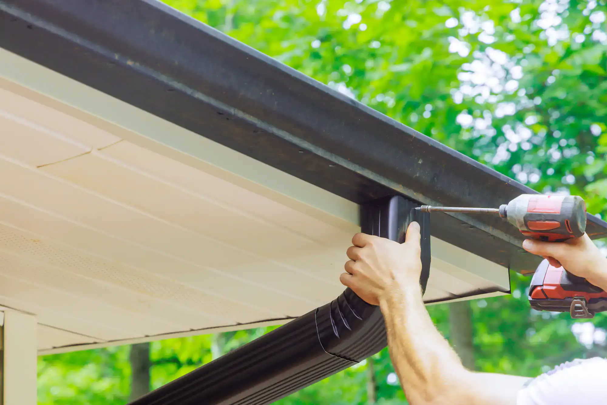 A person uses a power drill to attach a black gutter to the edge of a house roof, with green trees visible in the background.