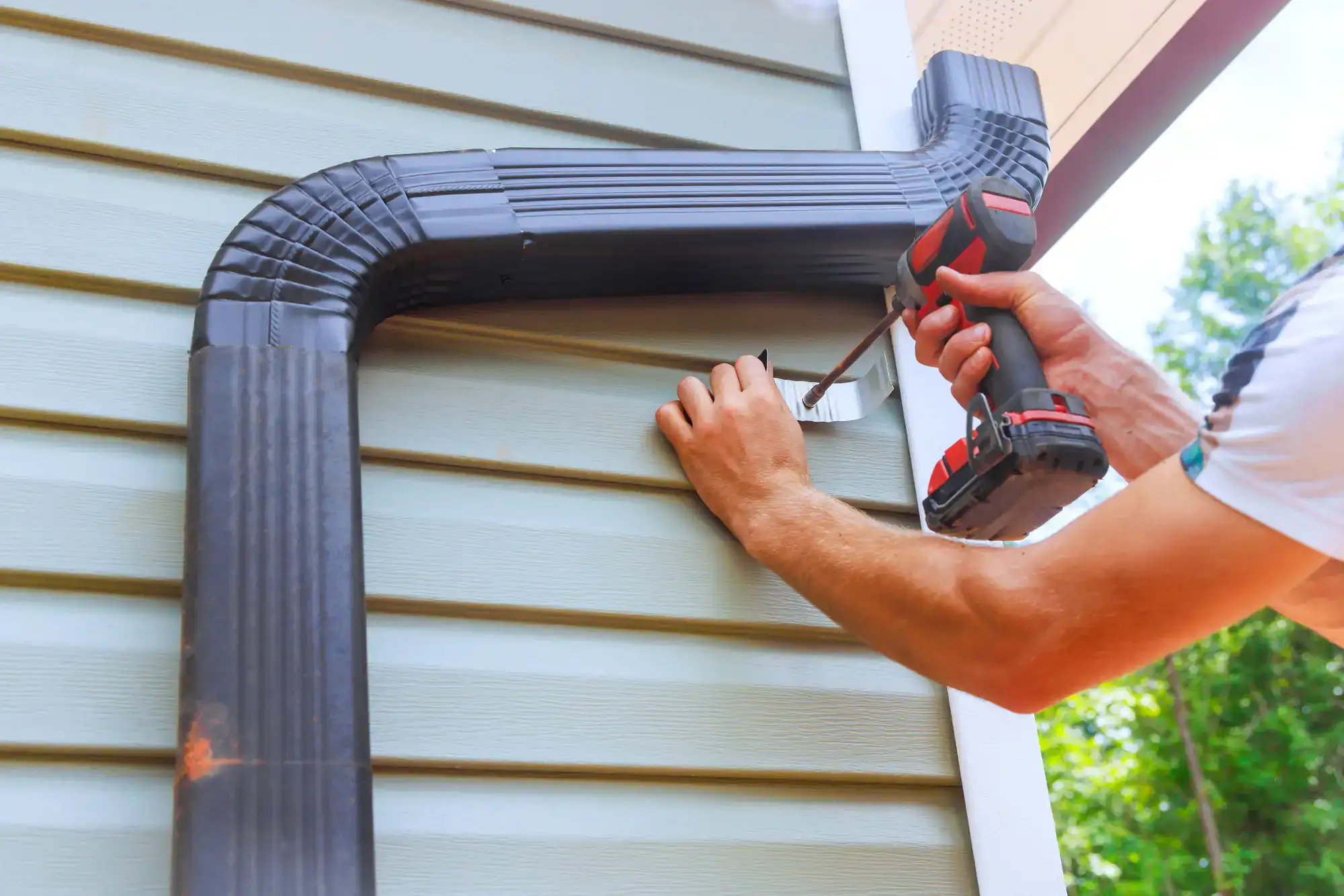 A person uses a power drill to secure a black metal downspout to the side of a house with light green siding on a sunny day.