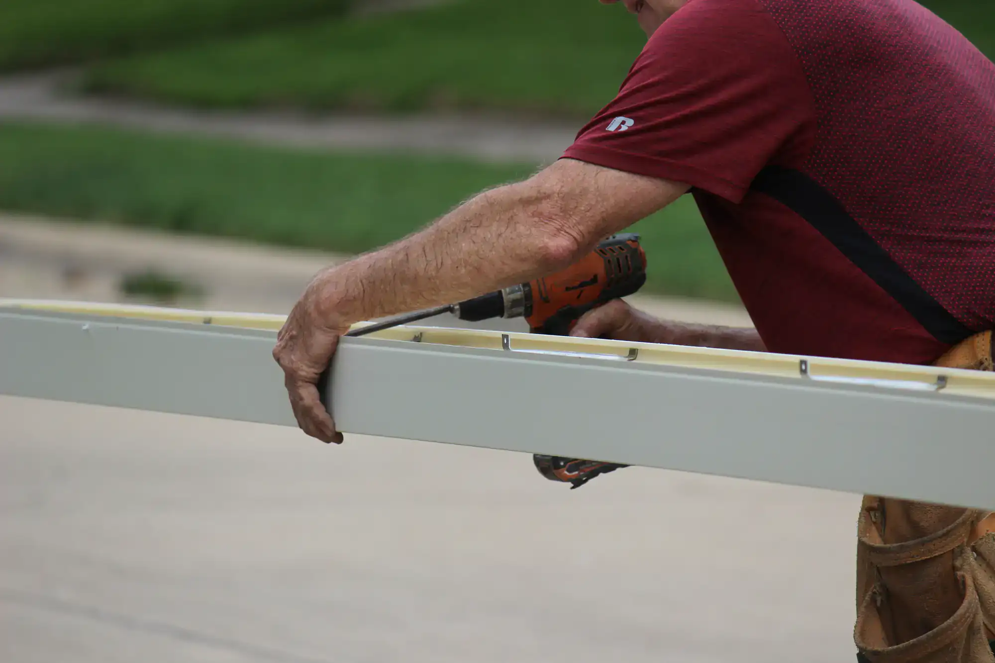A person wearing a red shirt uses a power drill to attach a screw into a long, white metal beam outdoors. Only the person's arms and torso are visible.