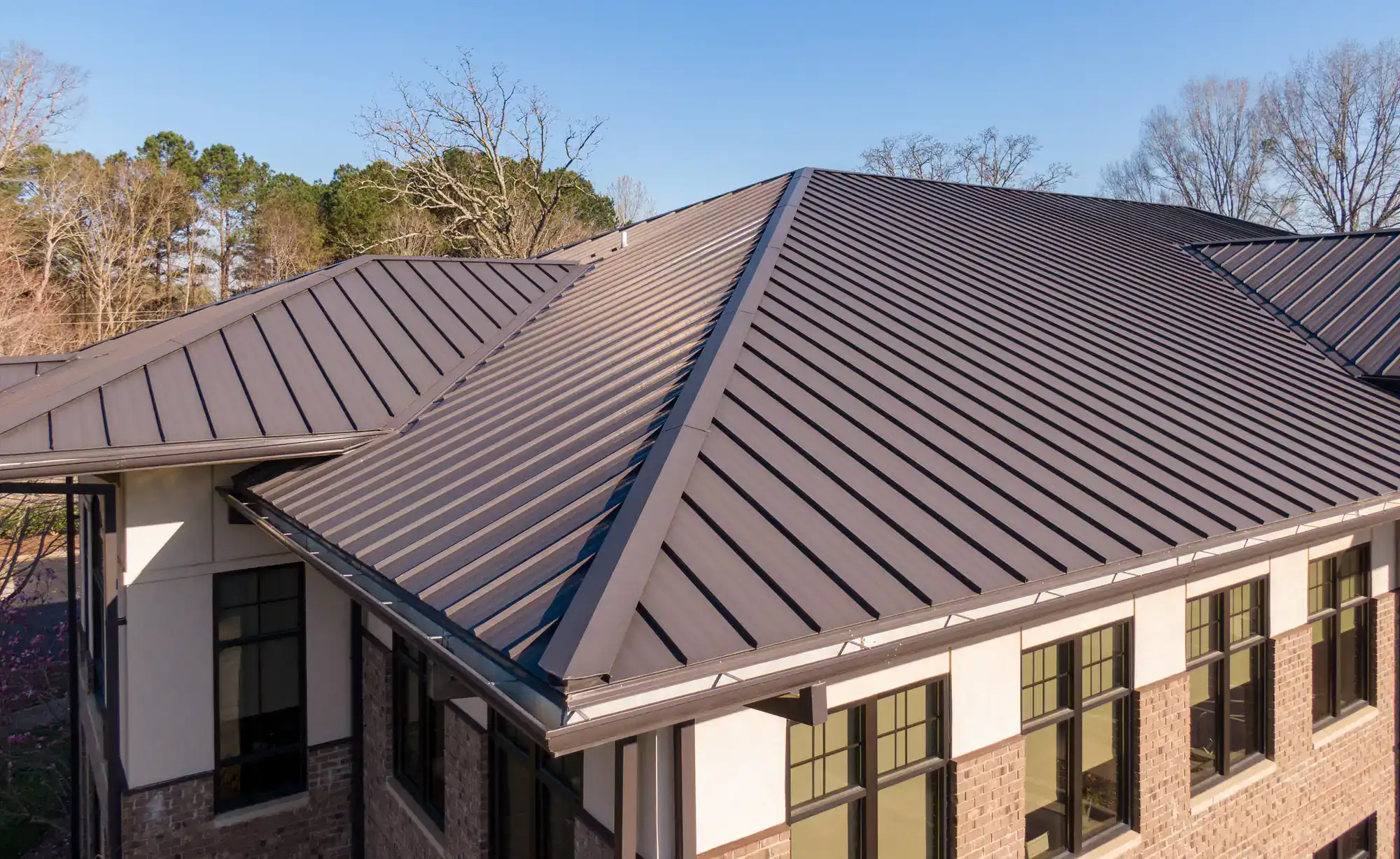 A close-up view of a modern building with a dark gray metal roof featuring standing seam panels, surrounded by trees and clear skies.