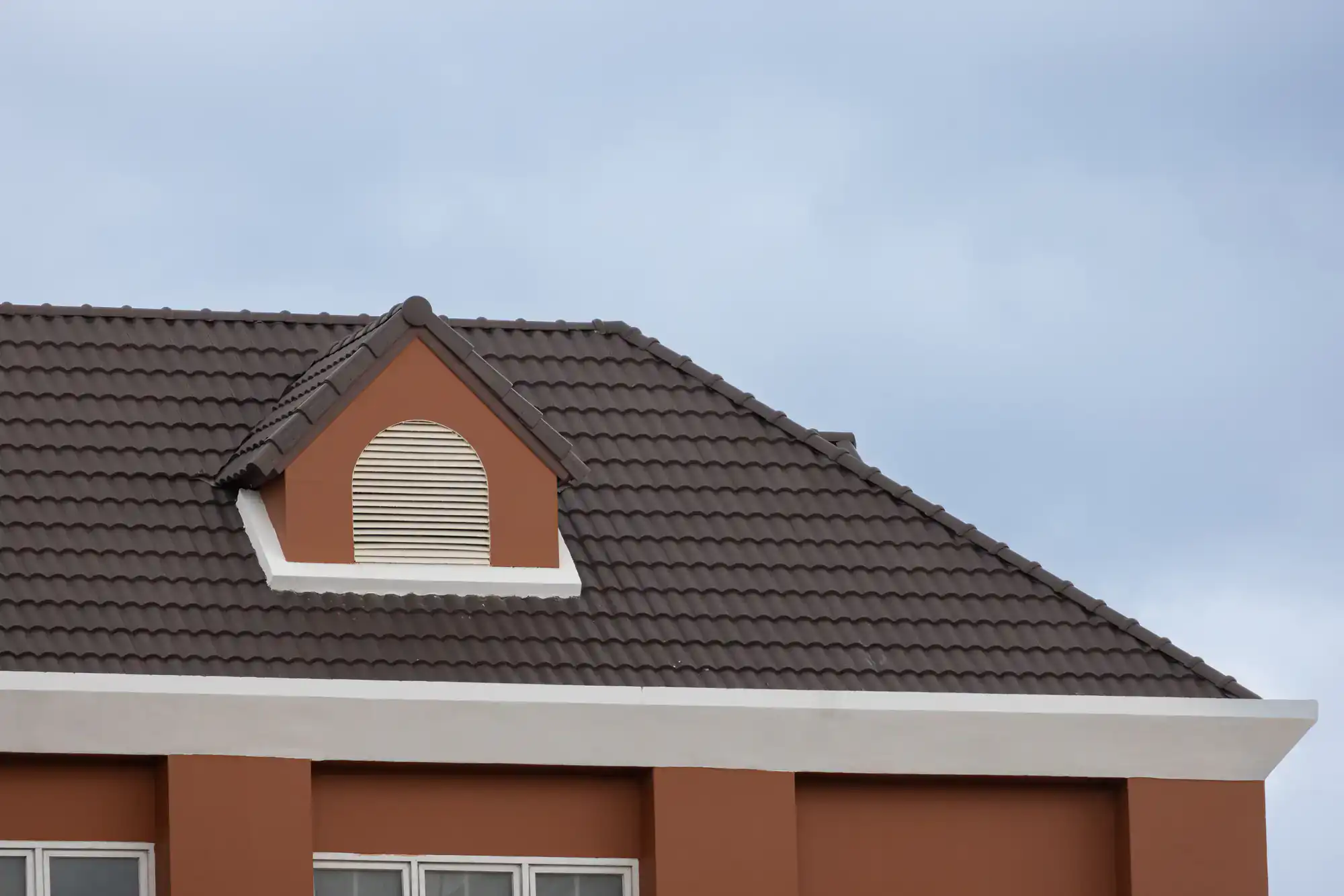 A close-up of a house roof with dark brown tiles features a small, arched dormer vent with cream-colored shutters. The building below has a white trim and light brown walls, set against a cloudy sky.