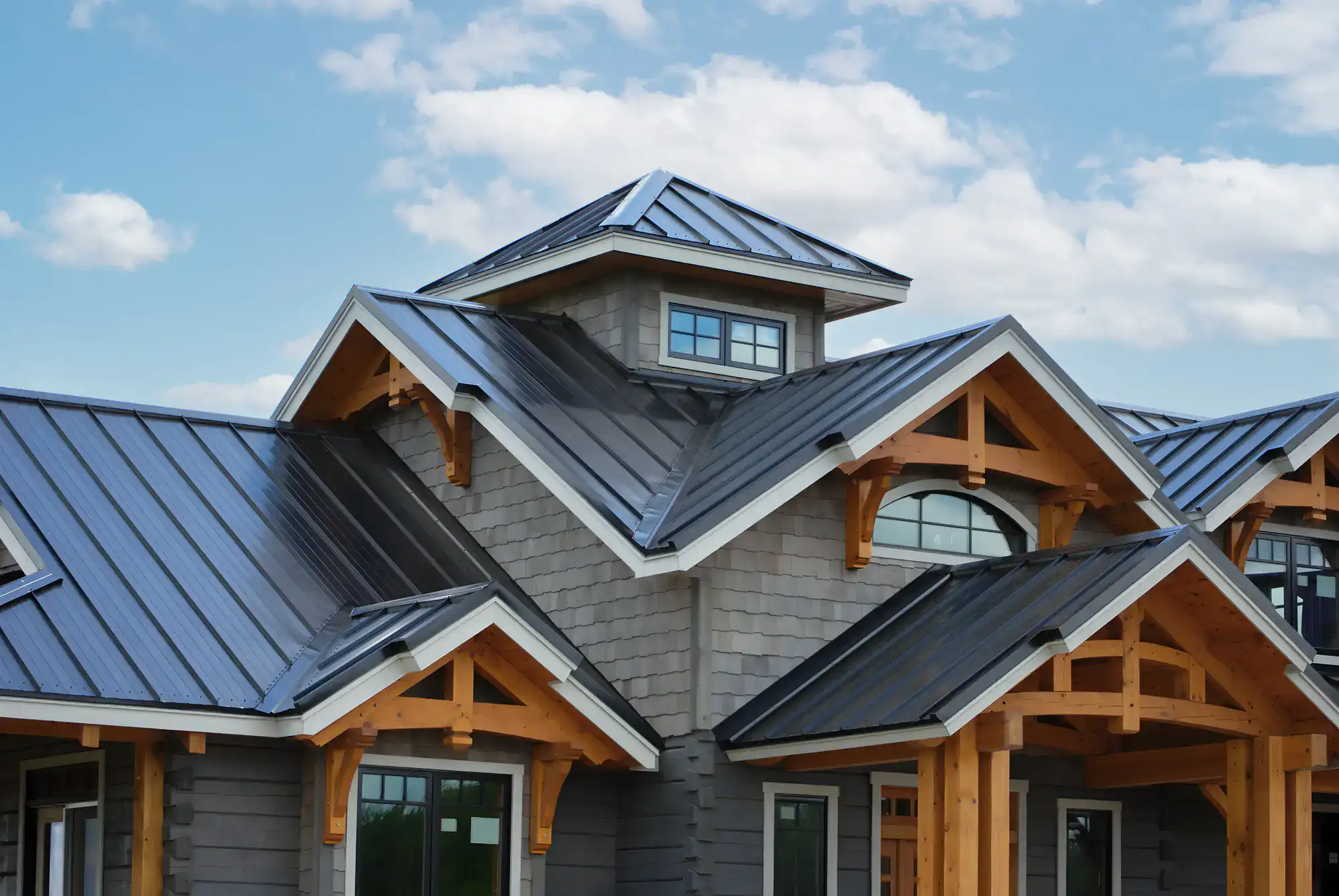 Modern house with gray metal roofing, wooden beams, and multiple peaked gables under a blue sky with scattered clouds. Large windows are visible below the rooflines.