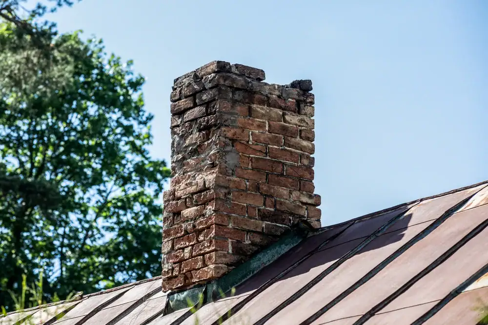 A weathered brick chimney with visible cracks rises from a sloped metal roof, surrounded by green trees and set against a clear blue sky.