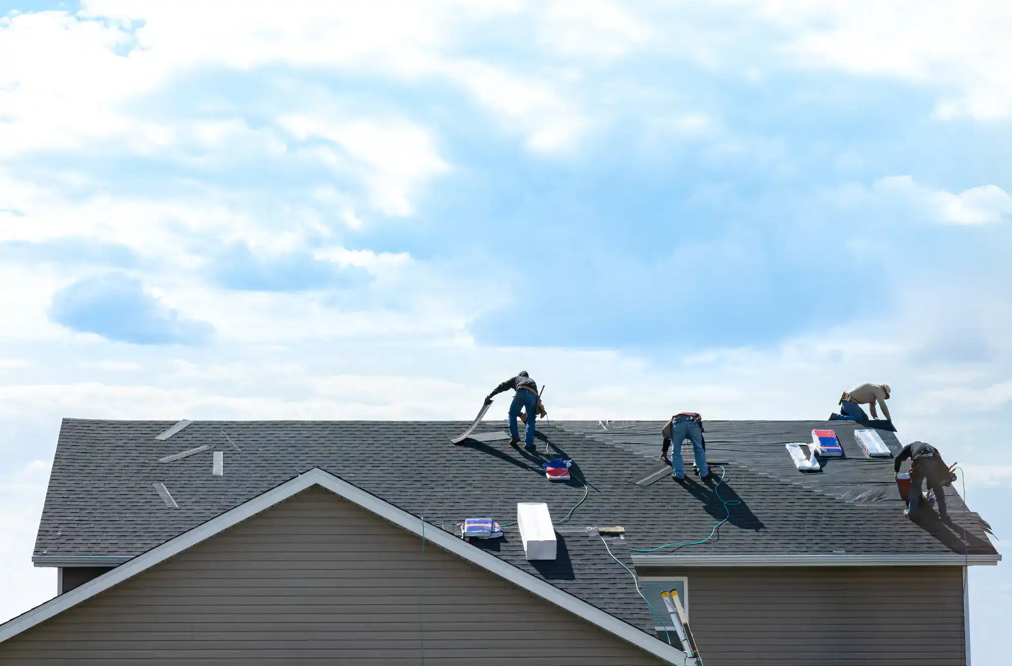 Four workers are installing or repairing shingles on a sloped residential roof under a bright, partly cloudy sky. Tools and roofing materials are scattered across the roof during this Home Improvement Suffolk County, NY project.