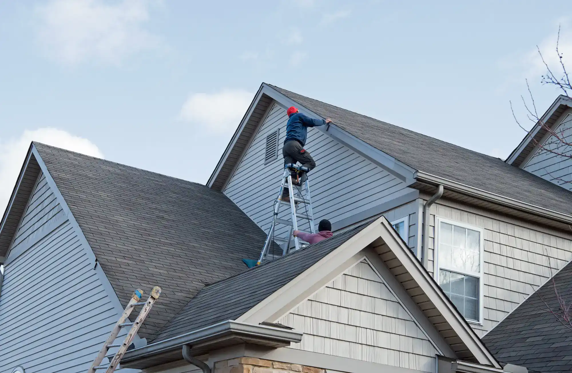 Two people work on the roof of a house; one stands on a ladder leaning against the roofline while the other sits on the roof. Another ladder is visible on the left side of the house. The sky is partly cloudy.