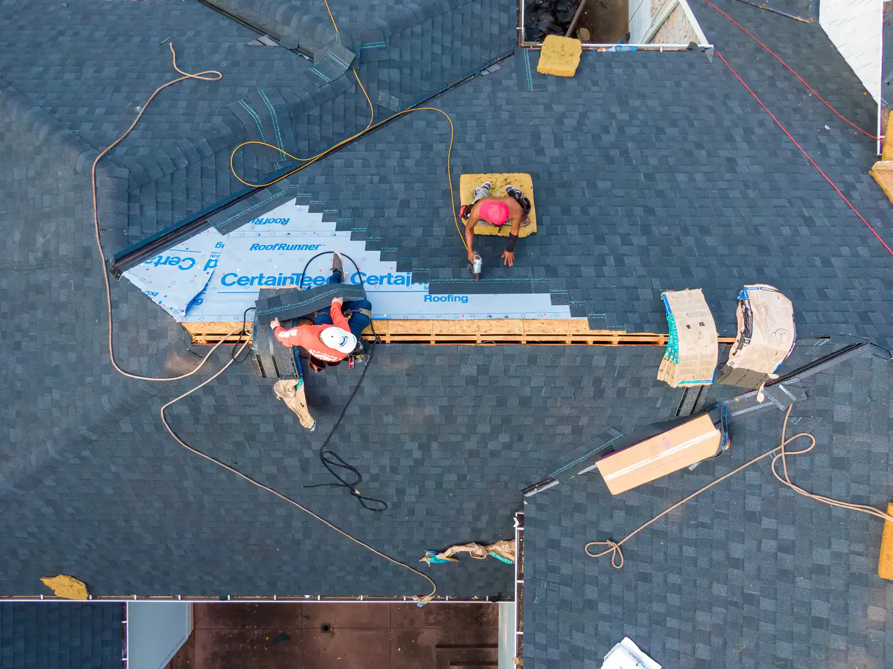 Three workers are installing shingles and roofing materials on a house roof. Viewed from above, they use tools and safety gear, with supplies and equipment scattered around the gray shingled roof.