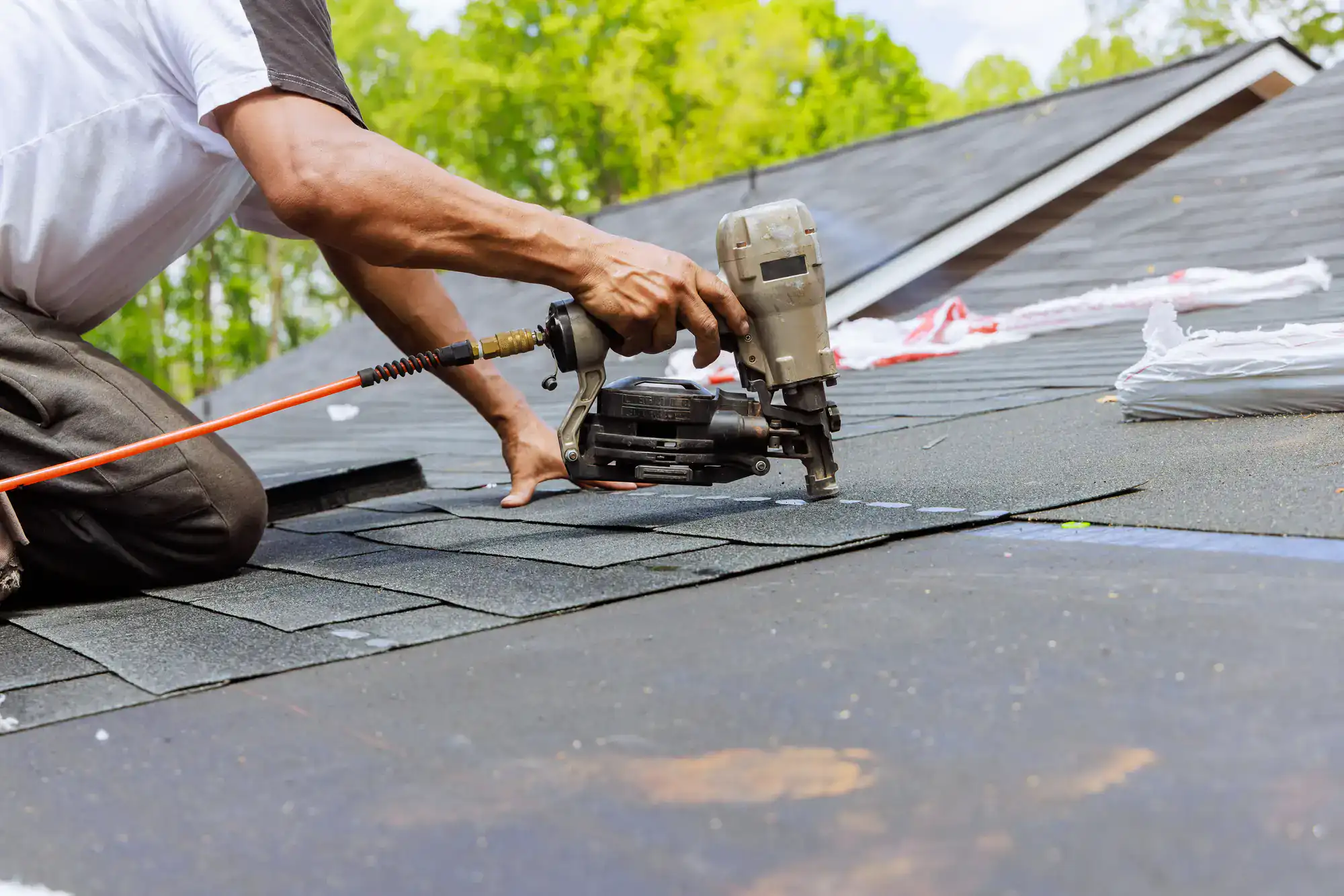 A person uses a nail gun to install asphalt shingles on a roof during a sunny day, with green trees visible in the background.