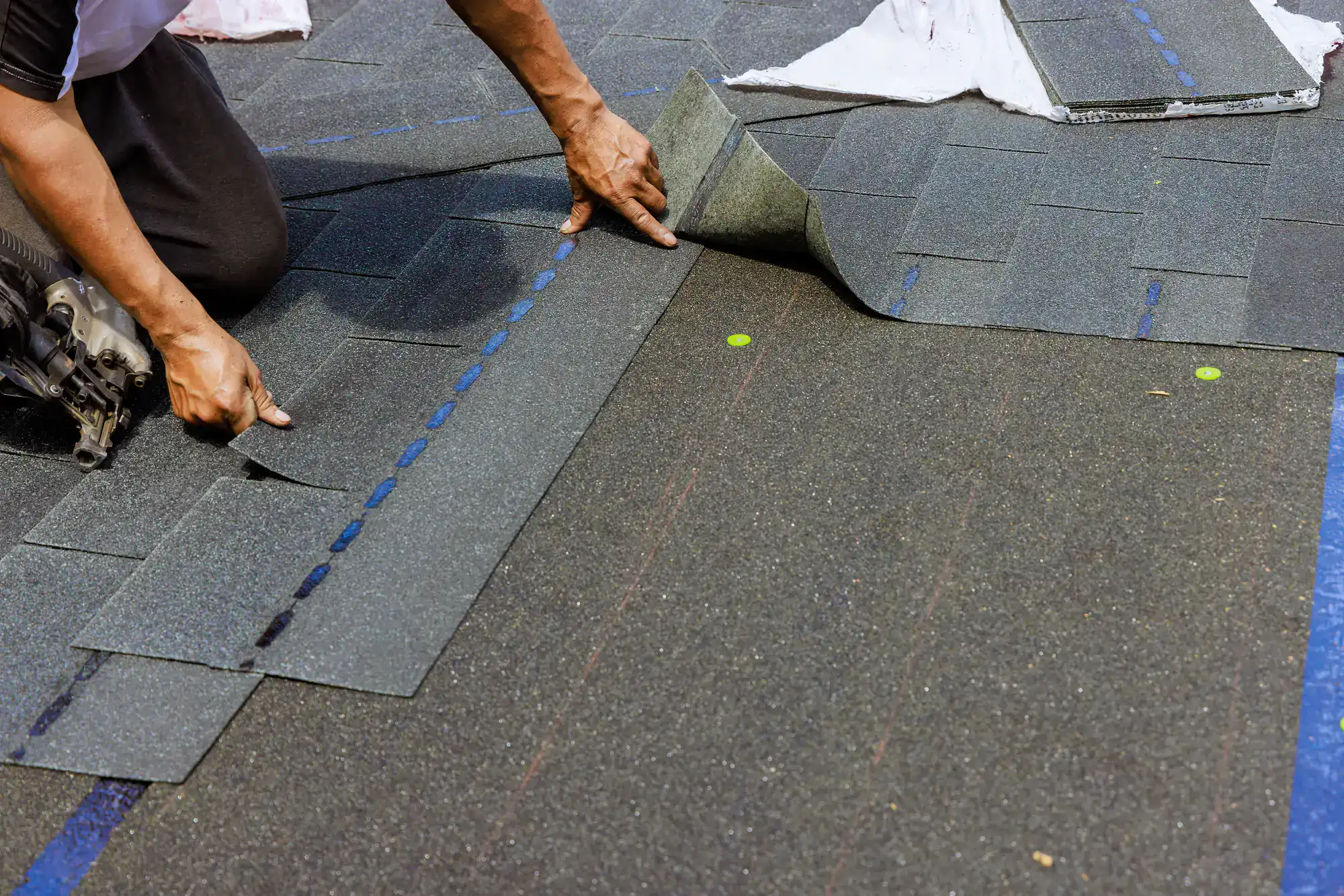 A person installs asphalt shingles on a roof, using both hands to position and secure the material in place. Tools and markings are visible on the surface.