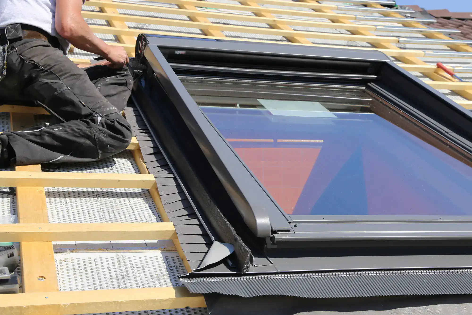A person installing or adjusting a skylight window on a sloped roof, surrounded by wooden battens and underlayment materials. The window frame is black and reflects the sky.