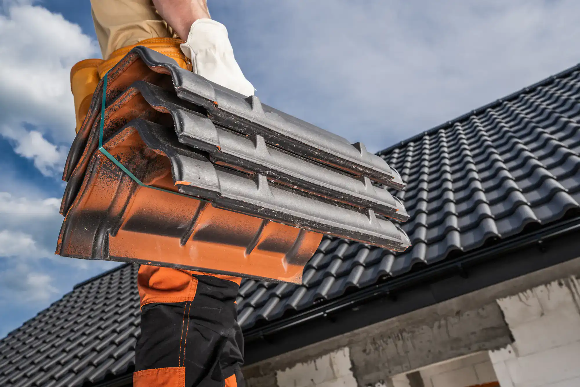 A worker wearing protective gloves carries several black roof tiles, standing near a house with a recently installed tiled roof under a blue sky with clouds.