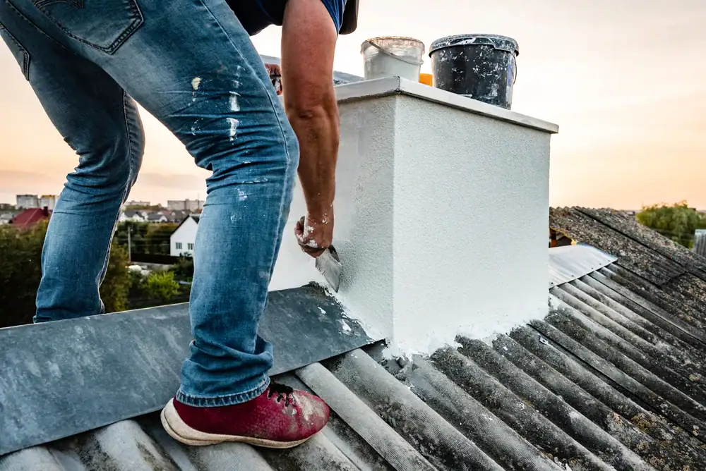 A person wearing jeans and red shoes is applying white plaster or paint to a rooftop chimney with a trowel. Buckets of materials are nearby, and the roof is made of corrugated metal sheets.