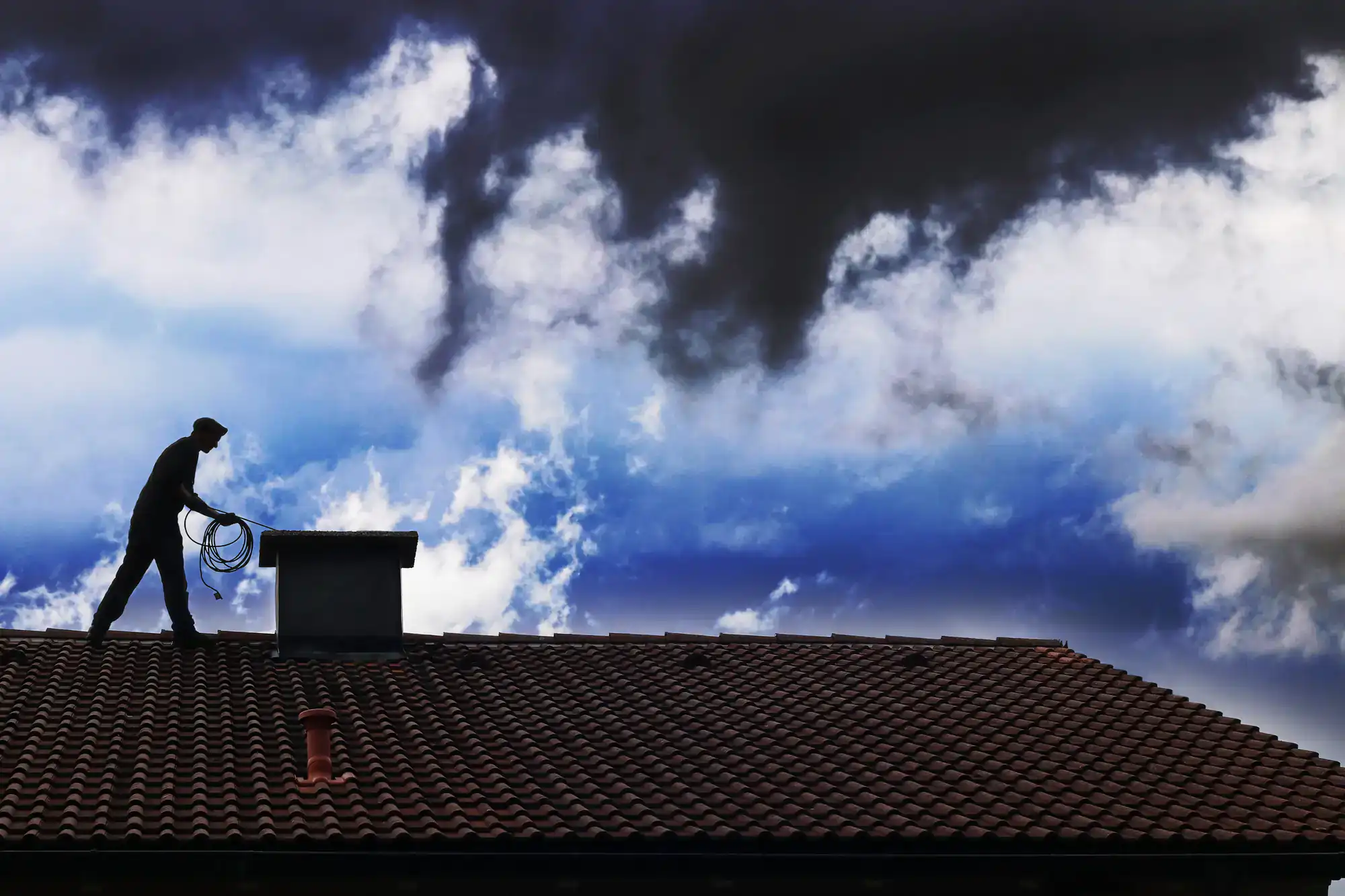 A person stands on a tiled roof holding coiled rope, silhouetted against a dramatic cloudy sky with patches of blue.
