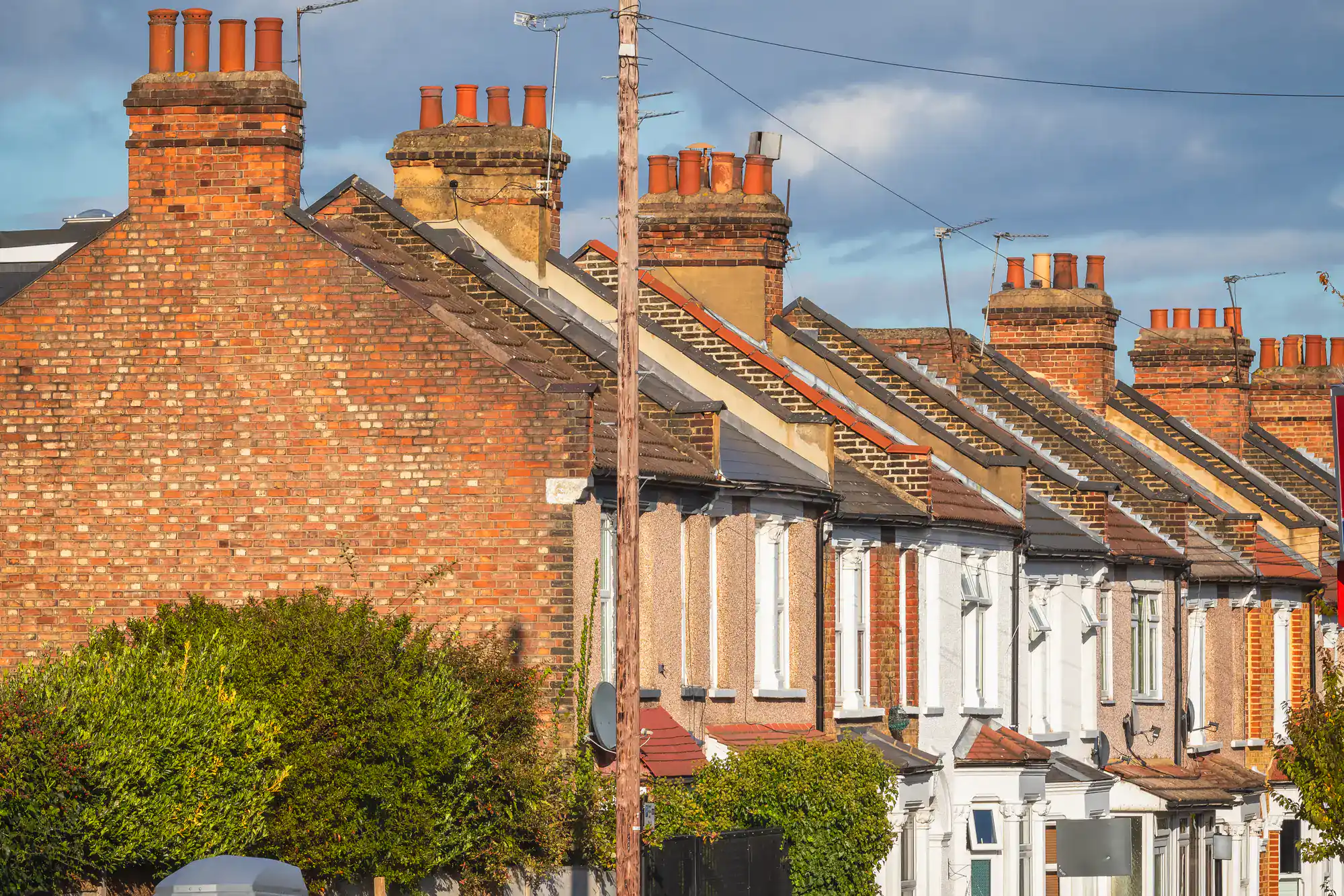 A row of traditional terraced houses with brick chimneys under a blue sky, sunlight highlighting the textures of the red and brown bricks; green shrubs line the front gardens.
