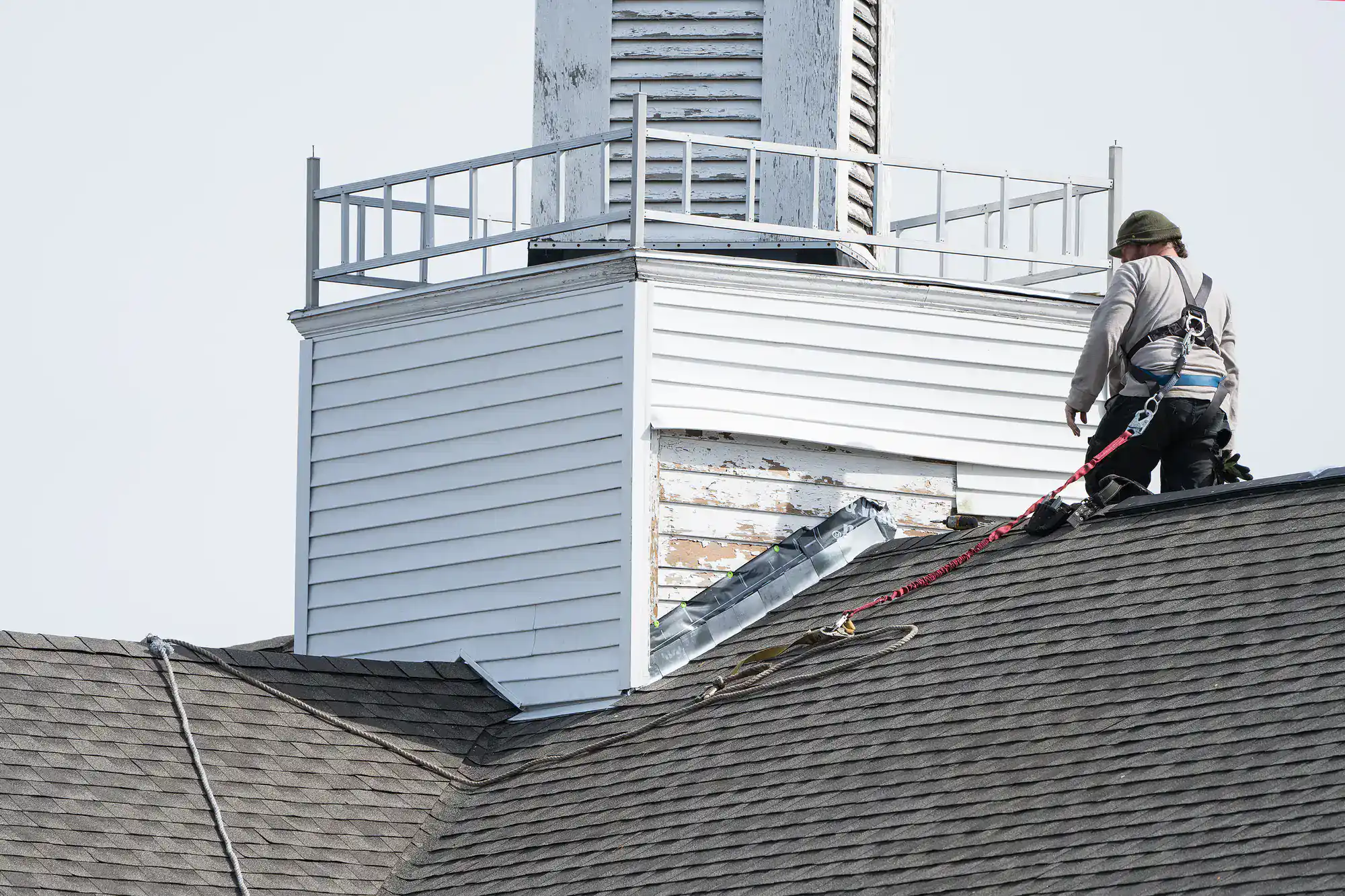 A worker in a safety harness repairs the siding of a steeple atop a shingled roof. The worker faces the structure, which shows signs of weathering and peeling paint. The sky is clear and bright.