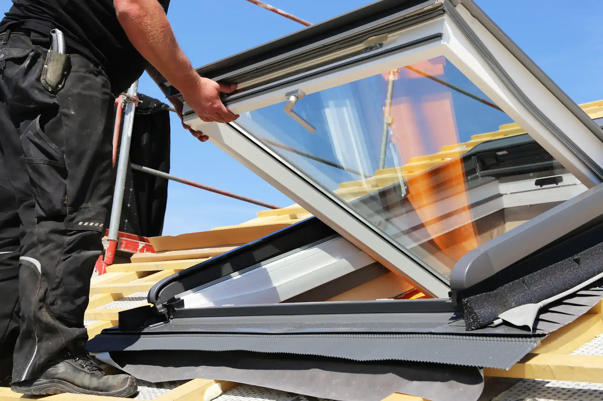 A person installs a skylight window on a sloped roof under a clear blue sky, surrounded by construction materials and tools.