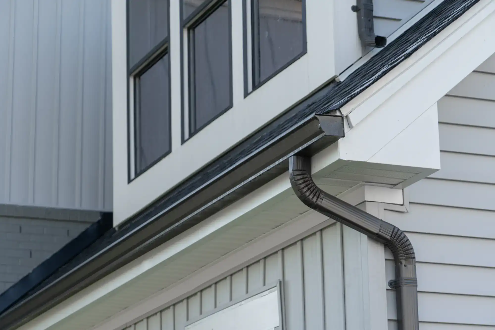 Close-up view of a house exterior, showing grey siding, white trim, black gutter and downspout system, and several large windows on the upper floor.