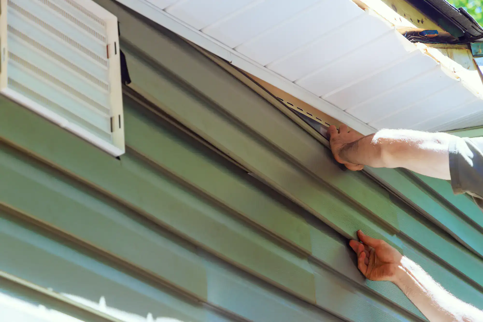 A person installs or repairs light green vinyl siding on the exterior wall of a house, using both hands to fit the siding panels under the white eaves.