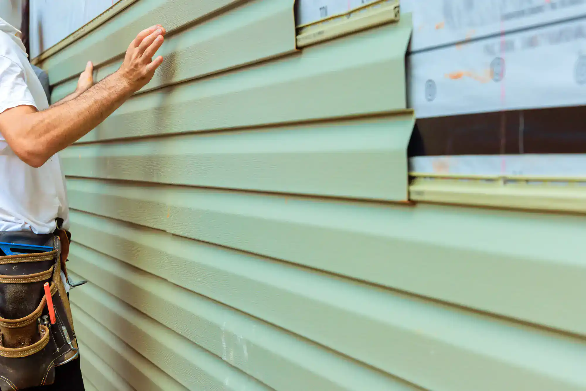A person wearing a tool belt installs light green vinyl siding on the exterior of a building, pressing the panels into place near a partially finished section.