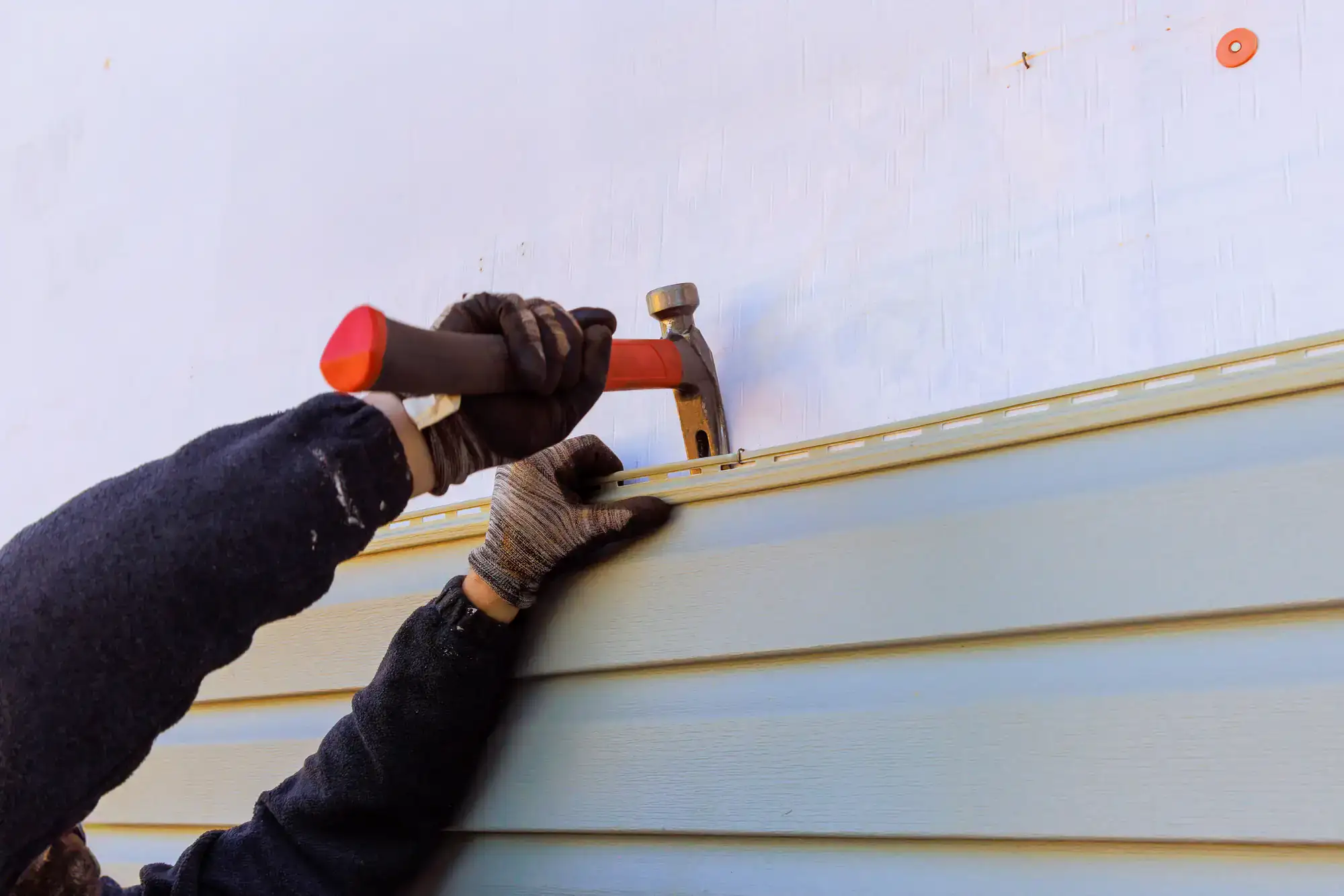 A person wearing gloves uses a hammer to nail light green vinyl siding onto the exterior wall of a building.