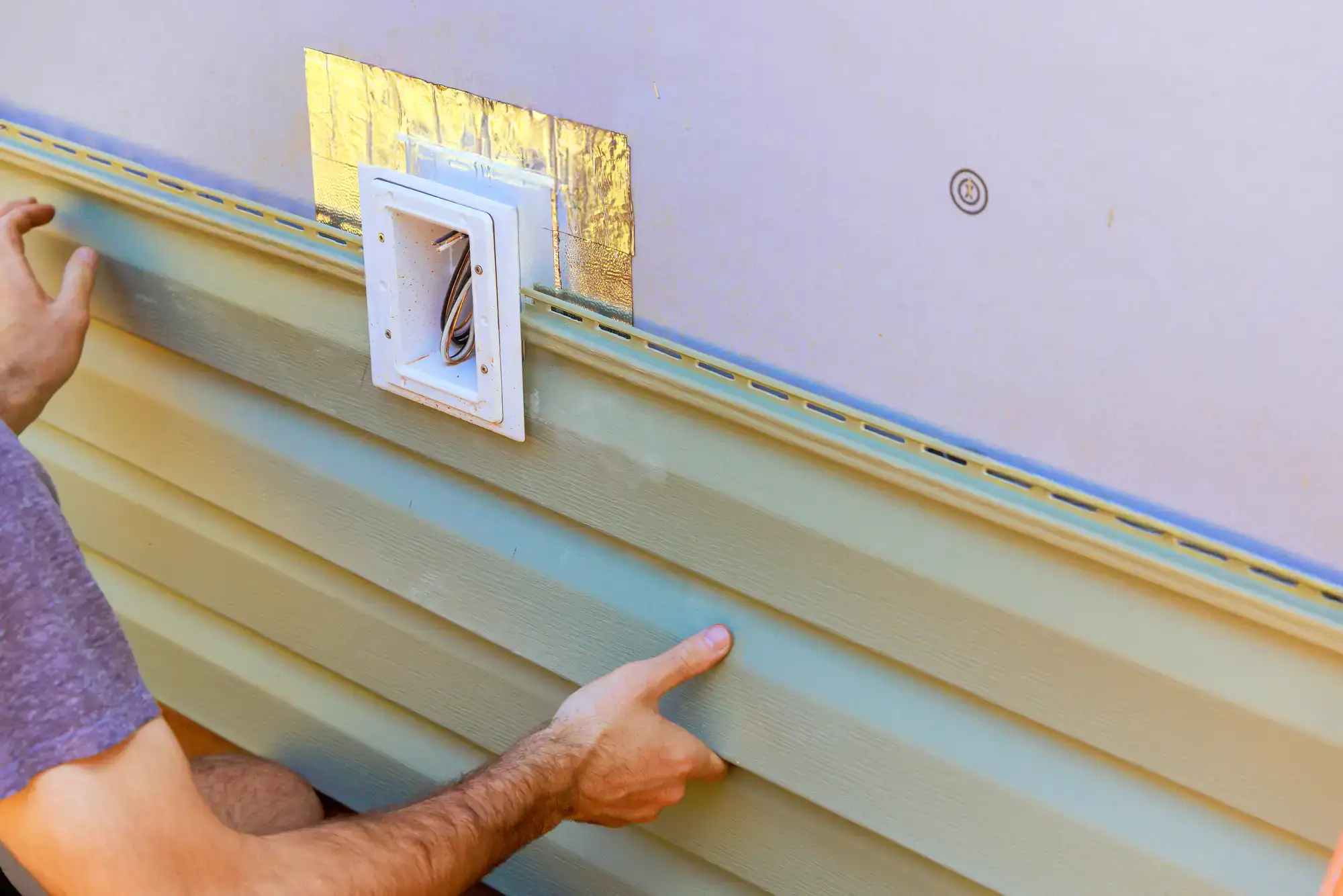 A person installs green vinyl siding on a house exterior, fitting the panel around a white utility vent on the wall.