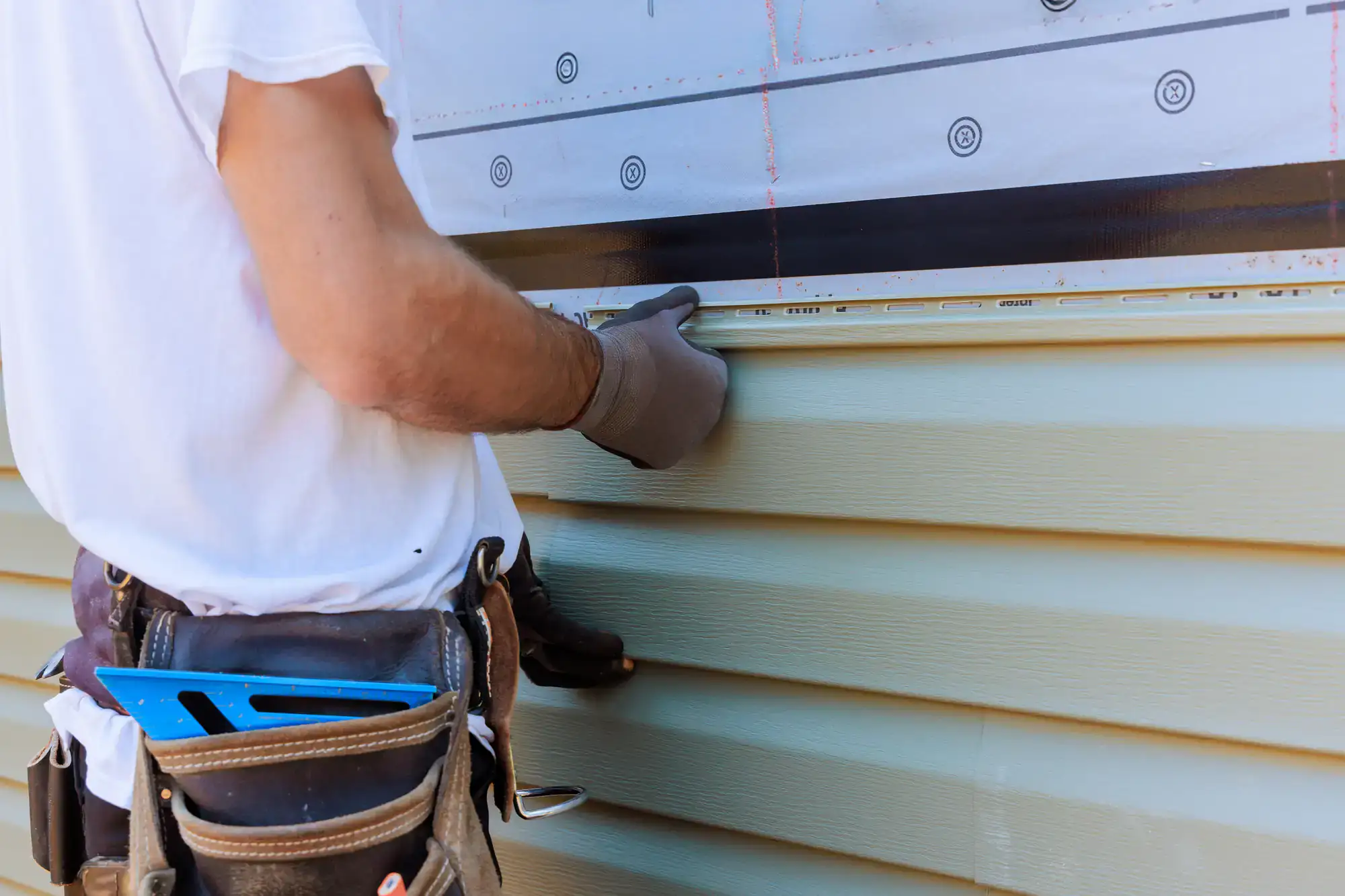 A construction worker wearing gloves installs light green vinyl siding on a house exterior, using a measuring tool and utility belt with tools.