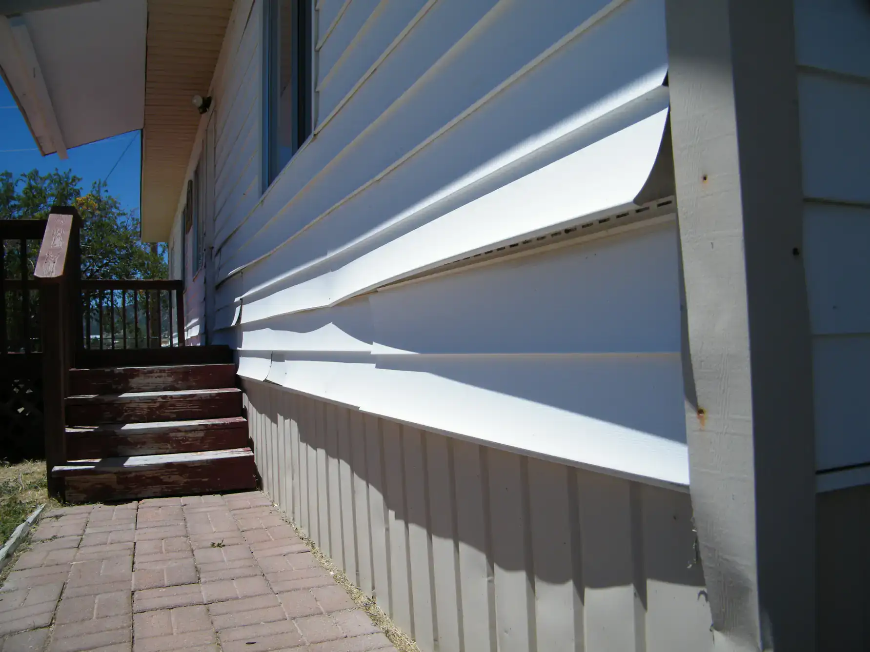 White vinyl siding on a house is warped and bent, creating uneven waves along the exterior wall near a set of wooden stairs and a paved walkway.