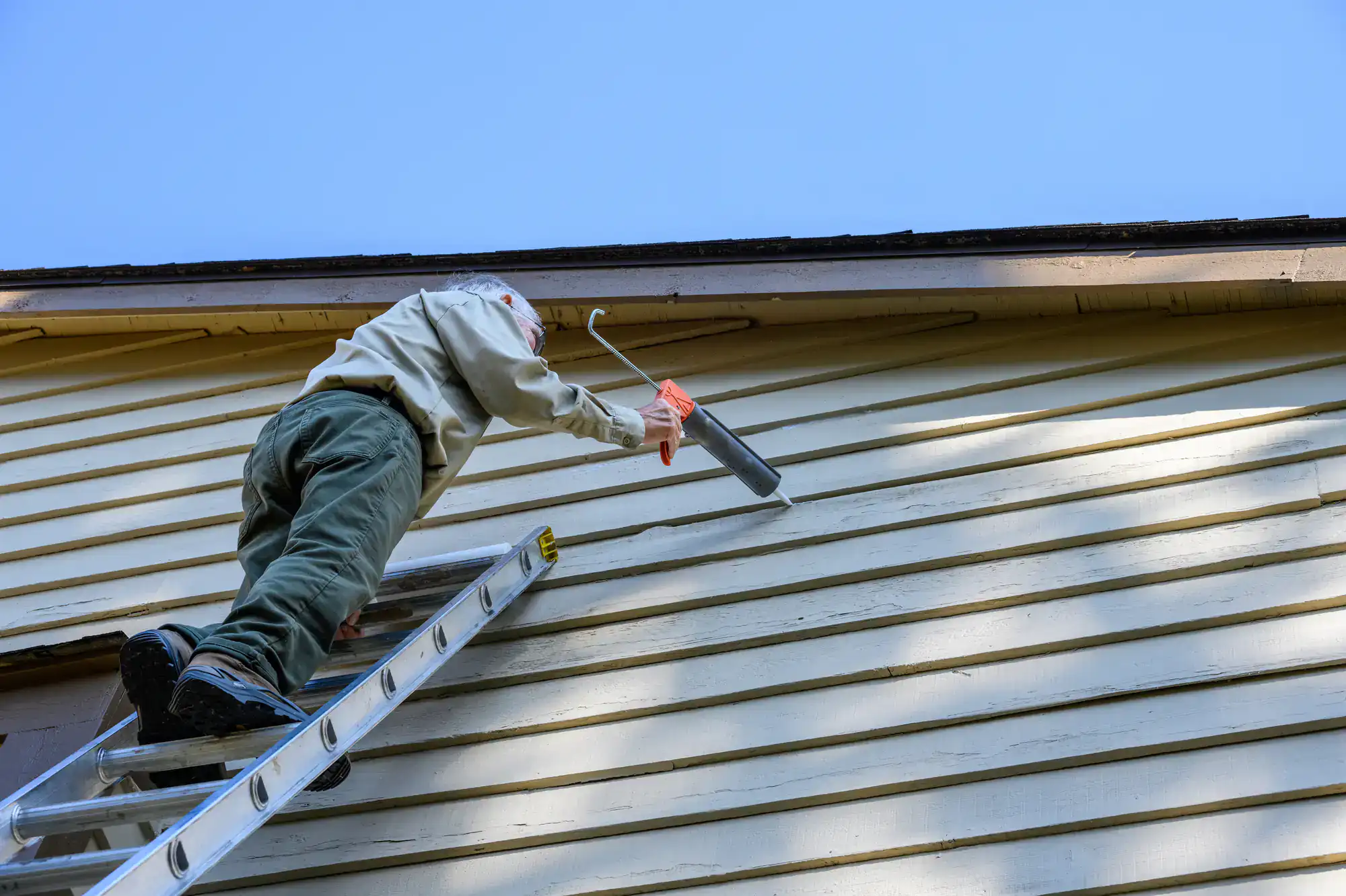 A person standing on a ladder applies caulk to the exterior siding of a house under a clear blue sky, likely performing home maintenance or repair work.