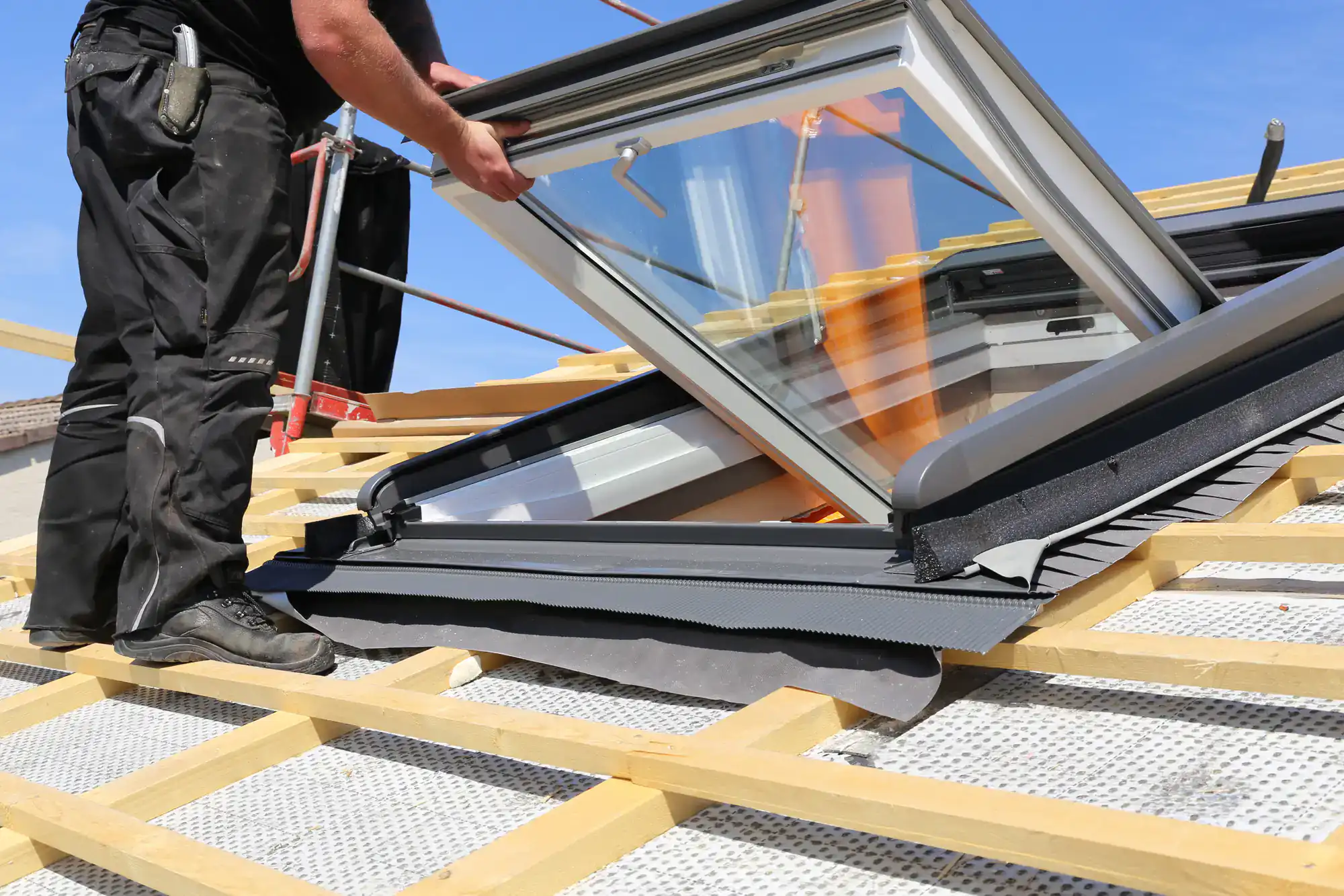 A worker installs a skylight window on a house roof, standing on wooden beams and underlayment on a sunny day.