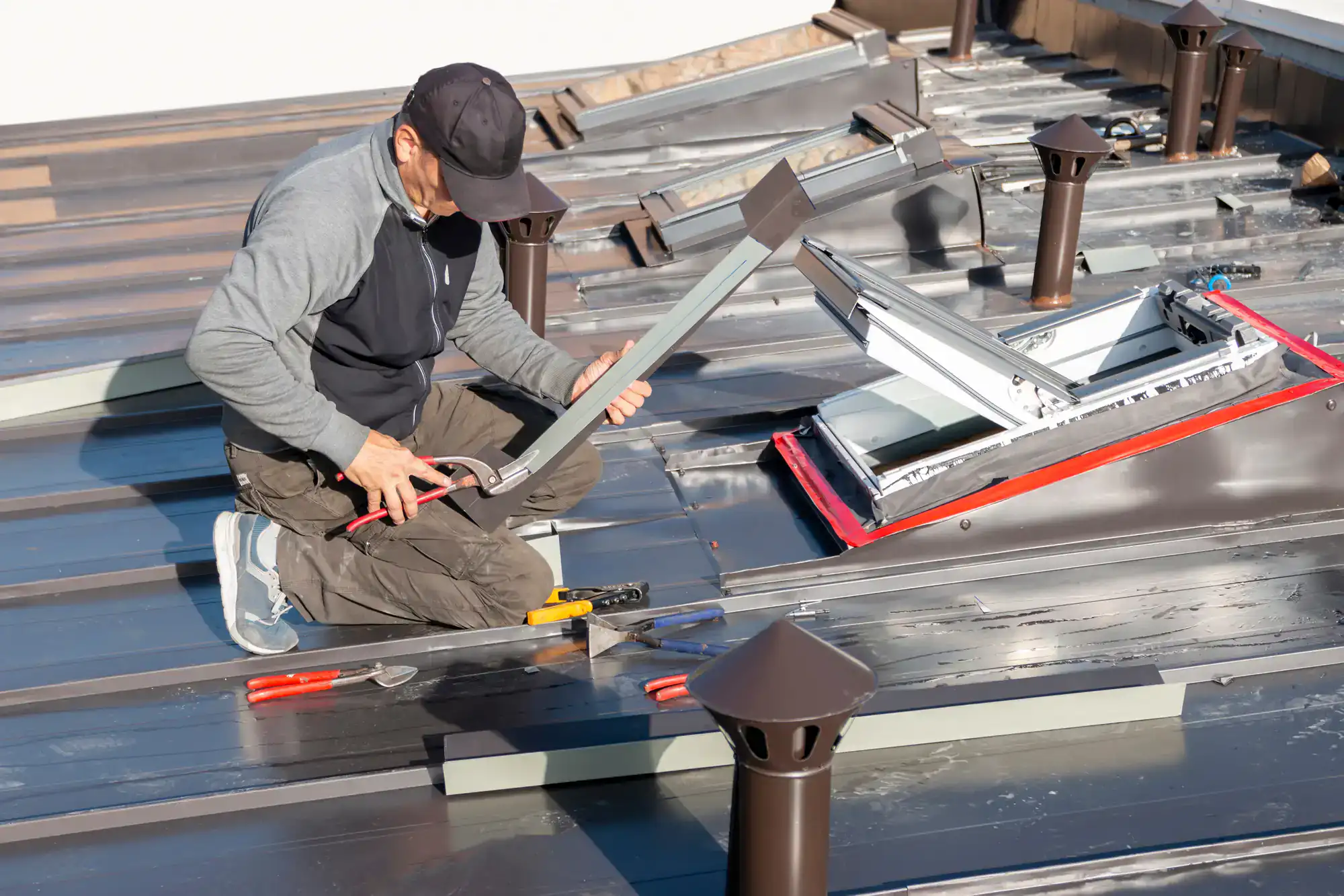 A man wearing a cap and work clothes is kneeling on a metal roof, using tools to install or repair a roof window. Various hand tools are scattered around him, and several roof vents are visible nearby.