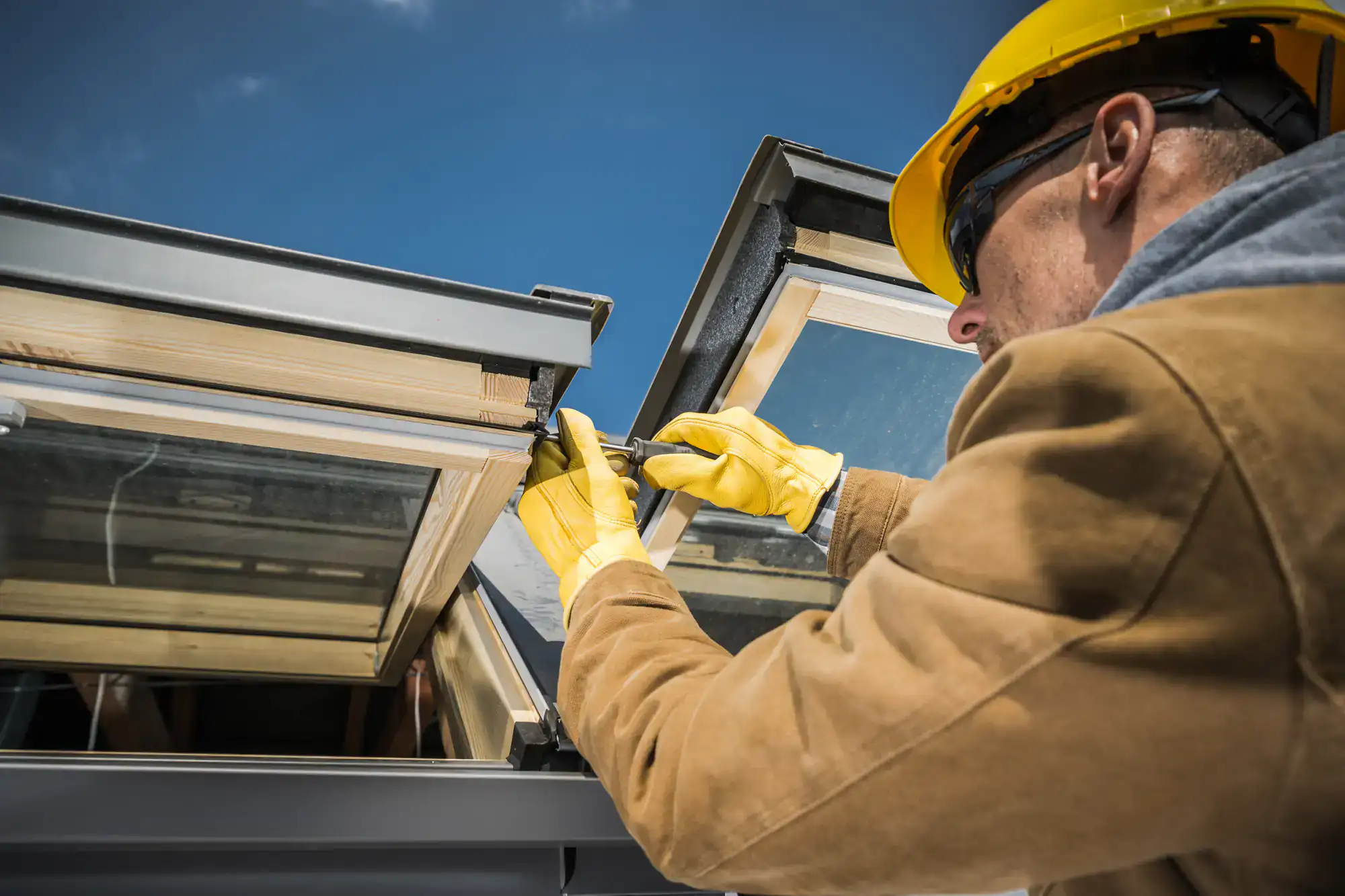 A construction worker wearing a yellow hard hat, safety glasses, and gloves installs or repairs a skylight window on a roof under a blue sky.