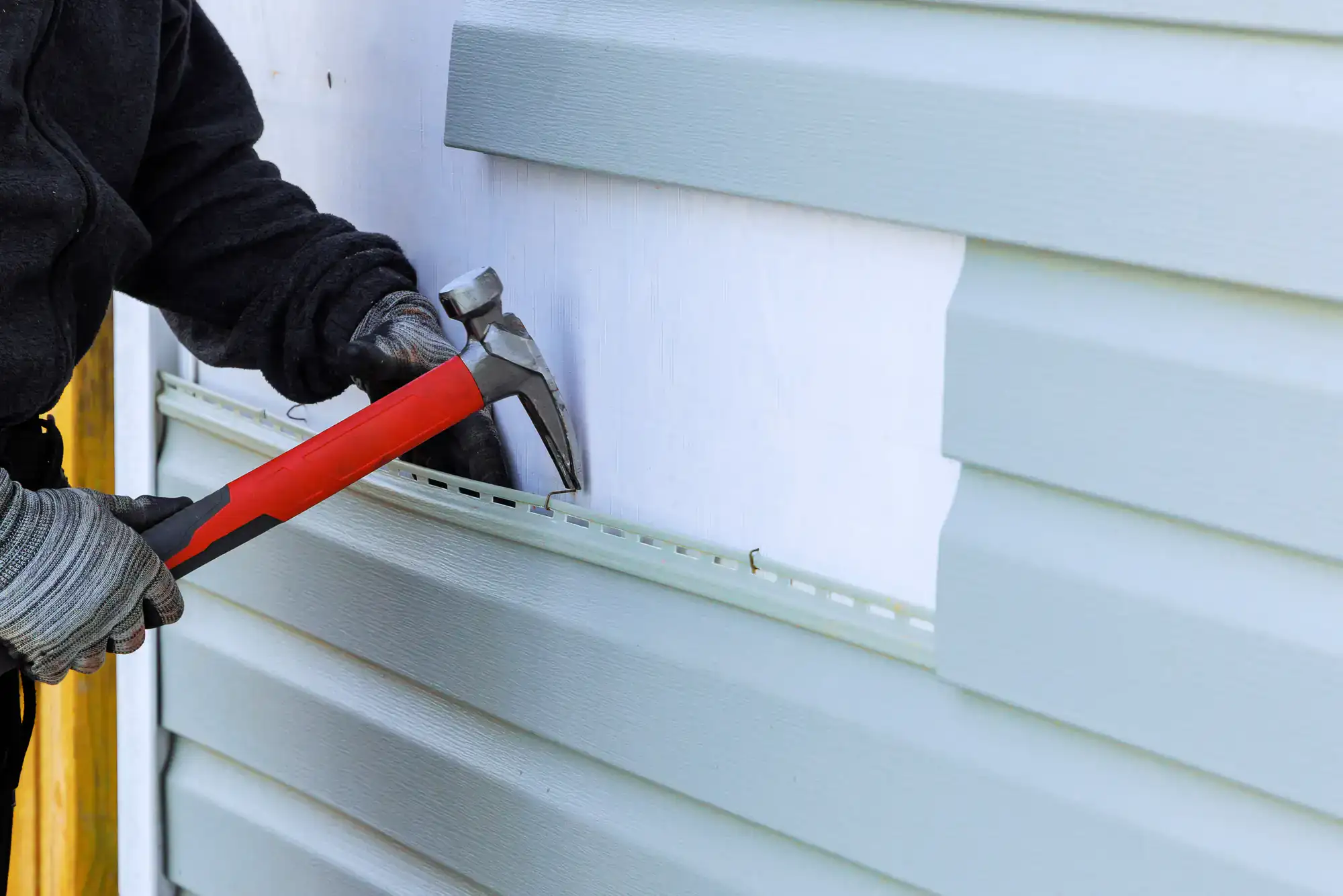 A person wearing gloves and a dark jacket uses a hammer to install light gray vinyl siding on the exterior wall of a building.
