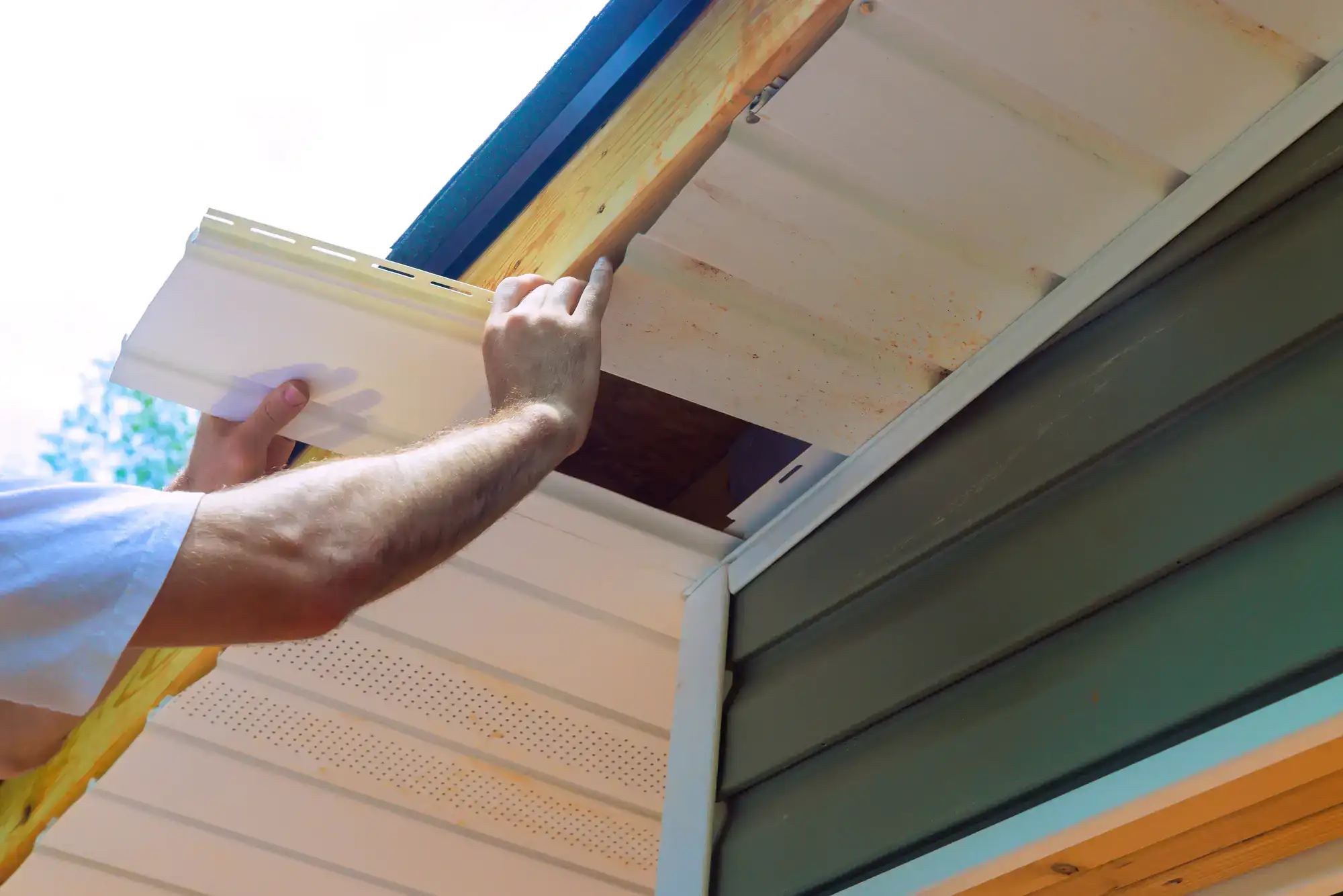 A person installs white vinyl soffit panels under the roof eaves of a house, using both hands to fit a panel in place. The house has green siding and exposed wooden beams.